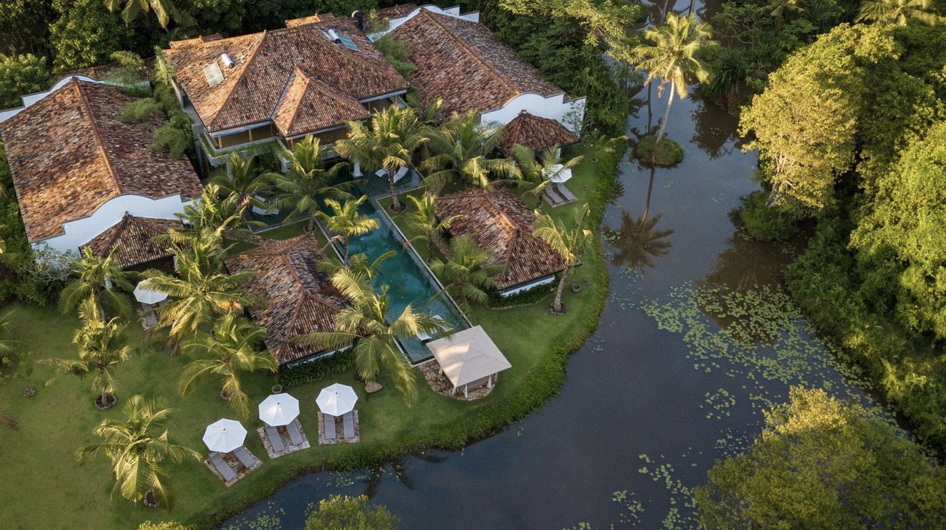 Aerial view of a tropical estate with multiple houses and pools surrounded by lush green trees and a water body with lily pads.