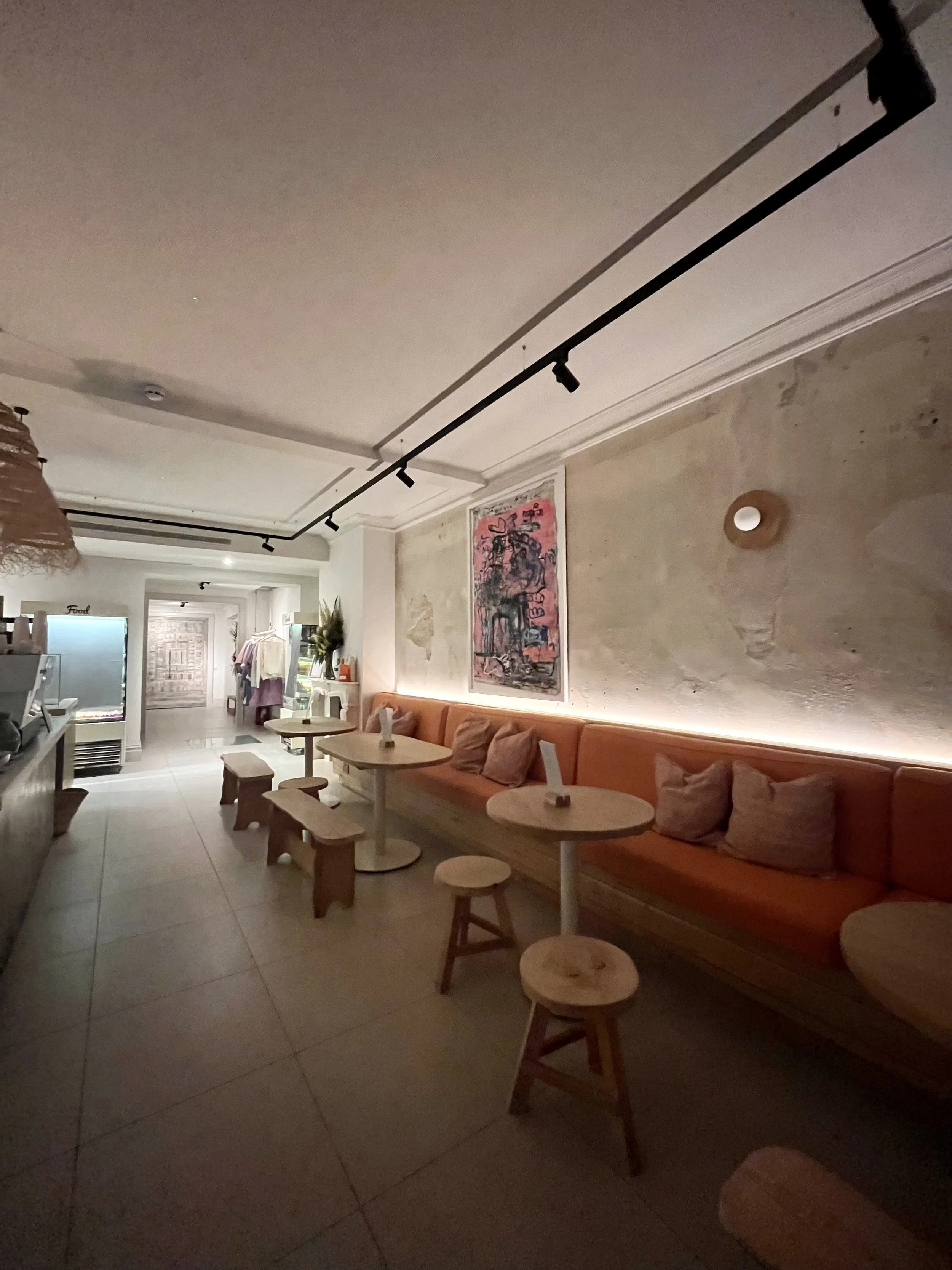 Interior of a cozy café with warm lighting, wooden benches with cushions, small round tables with stools, a wall art poster, and a display shelf with clothing and plants in the background.