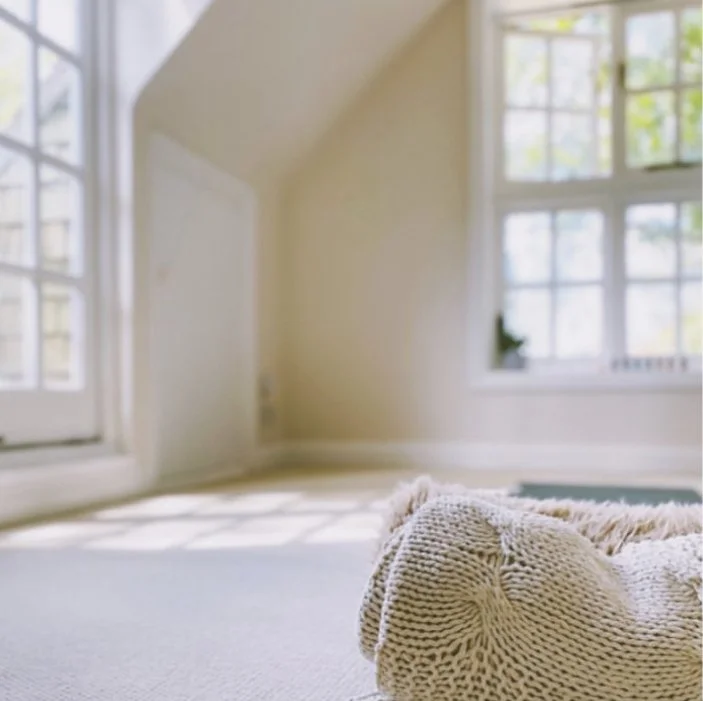 Empty room with large windows letting in sunlight and a cozy beige blanket or cushion on the floor.