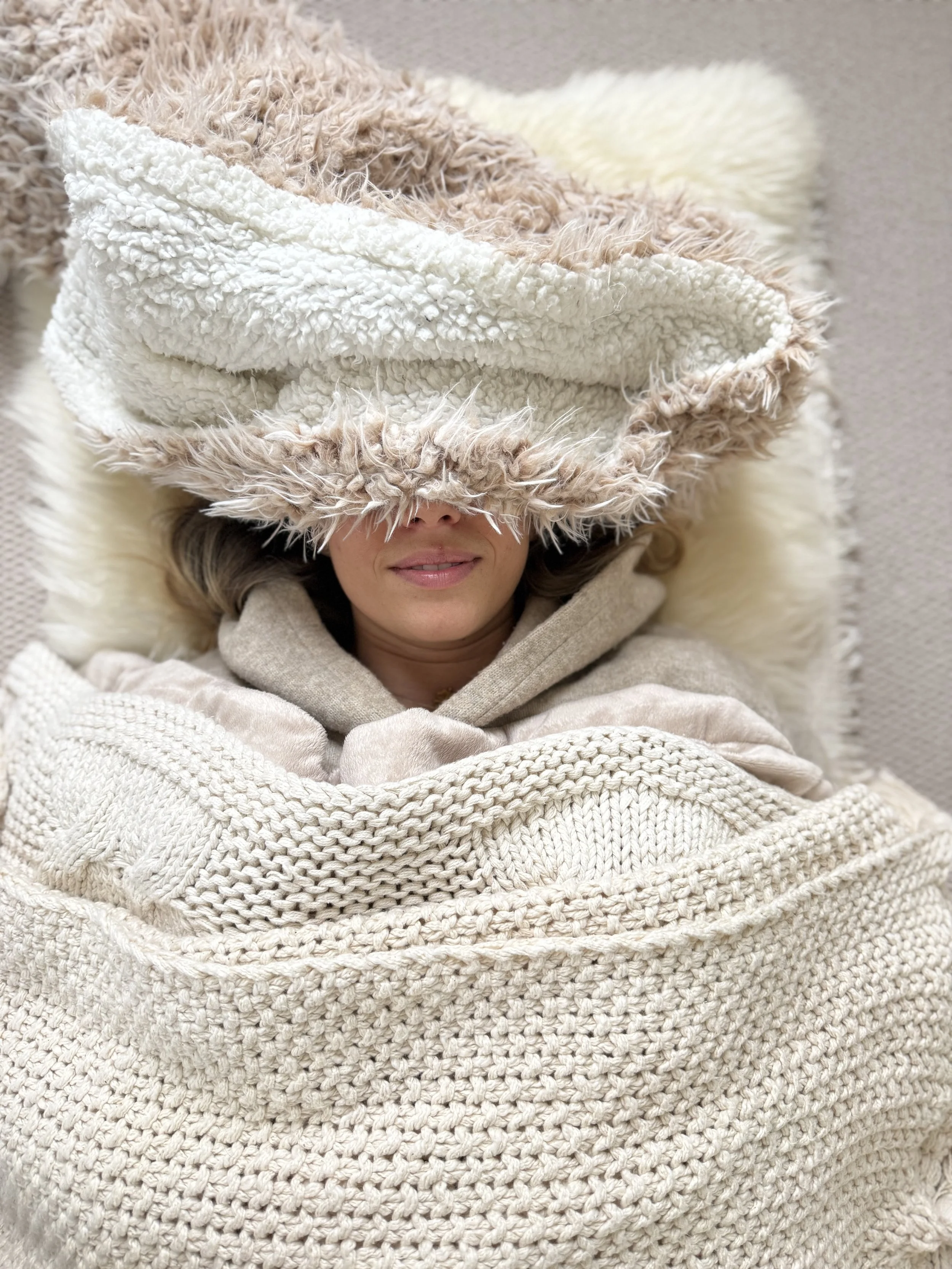 A woman lying down on a textured knit blanket, wearing a fluffy hat with a cream-colored fleece lining, and a cozy beige hoodie.