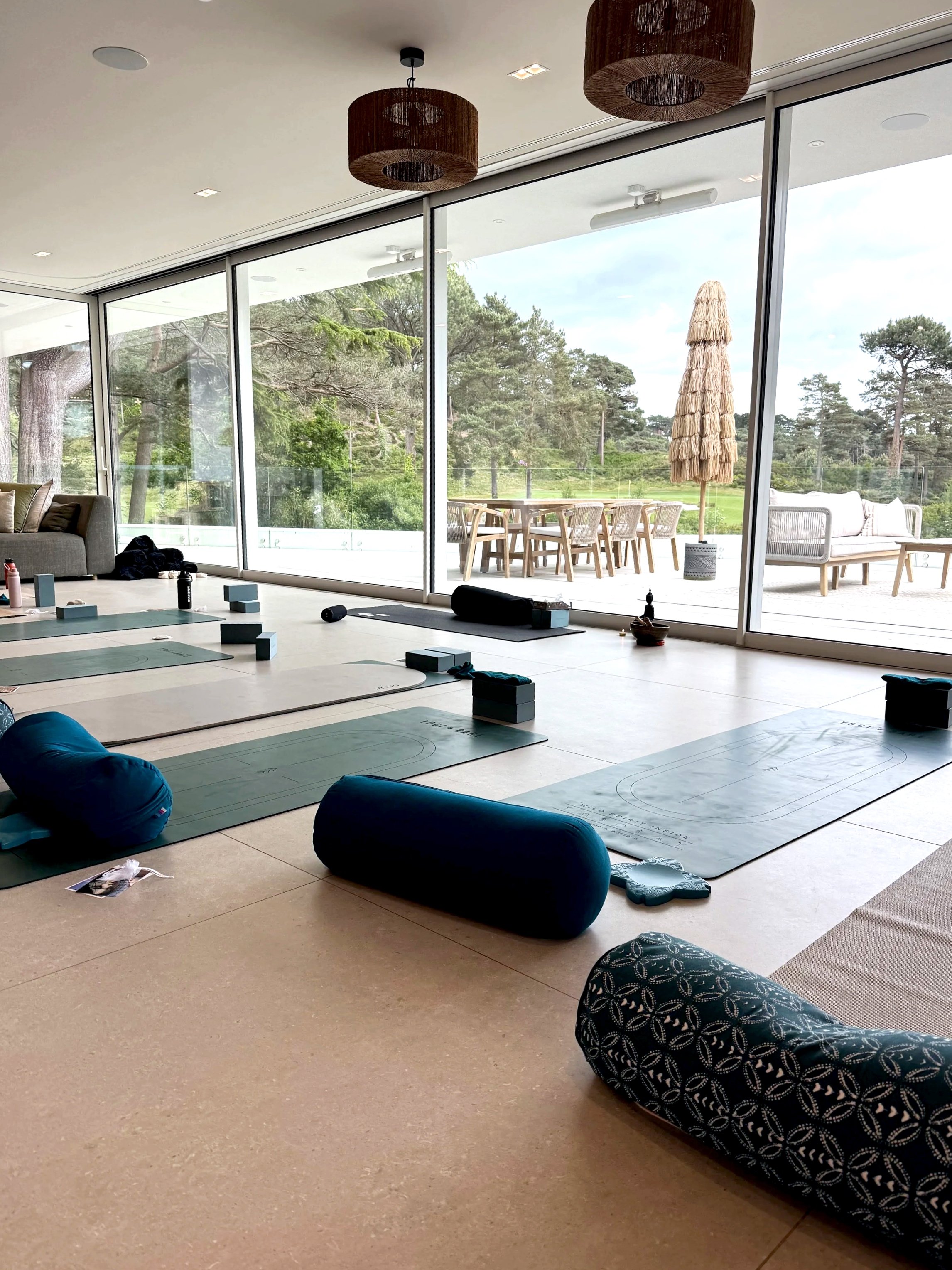 Indoor space with yoga mats, bolsters, and blocks arranged on a light-colored floor, next to large glass sliding doors leading to a patio with outdoor furniture and a thatched umbrella. Outside view shows trees and a green landscape.