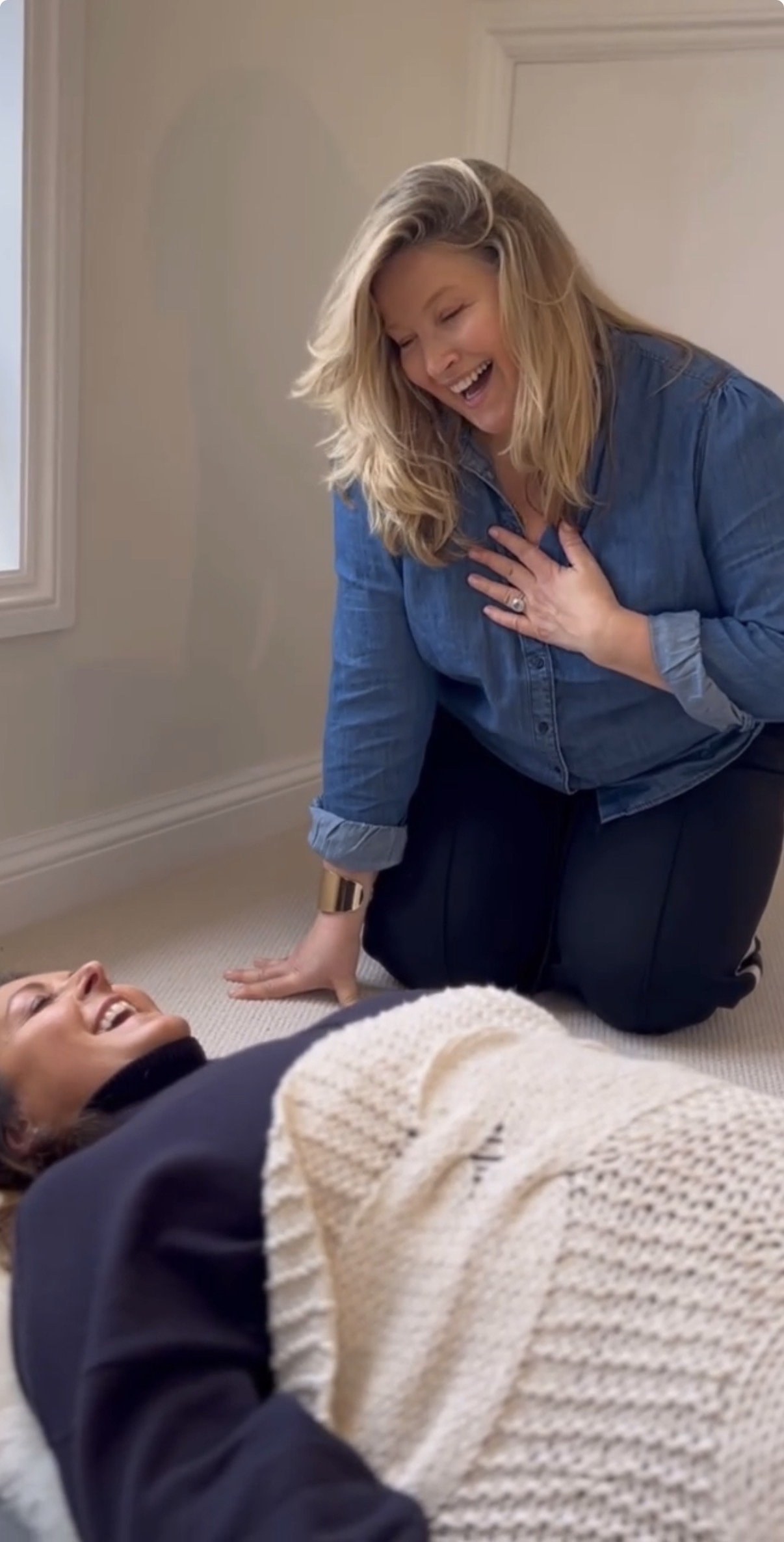 Two women appear to be laughing and enjoying each other's company in a warmly lit room with cream-colored walls. One woman is lying on the floor, while the other kneels beside her, touching her chest in a caring manner. The scene conveys a moment of joy or connection.