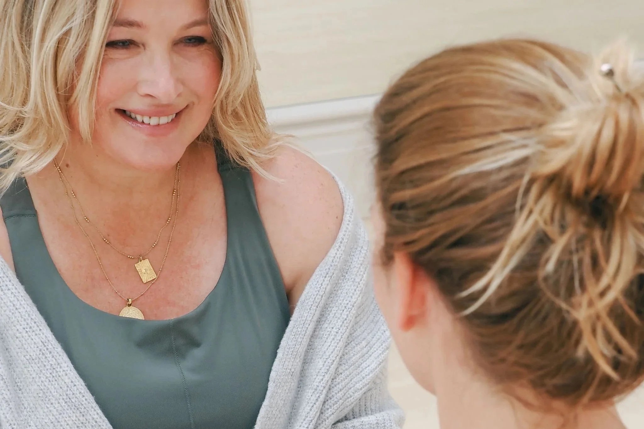 A smiling woman with blonde hair and a tan complexion sits opposite another person with red hair. She is wearing layered gold necklaces and a gray top with a white cardigan.