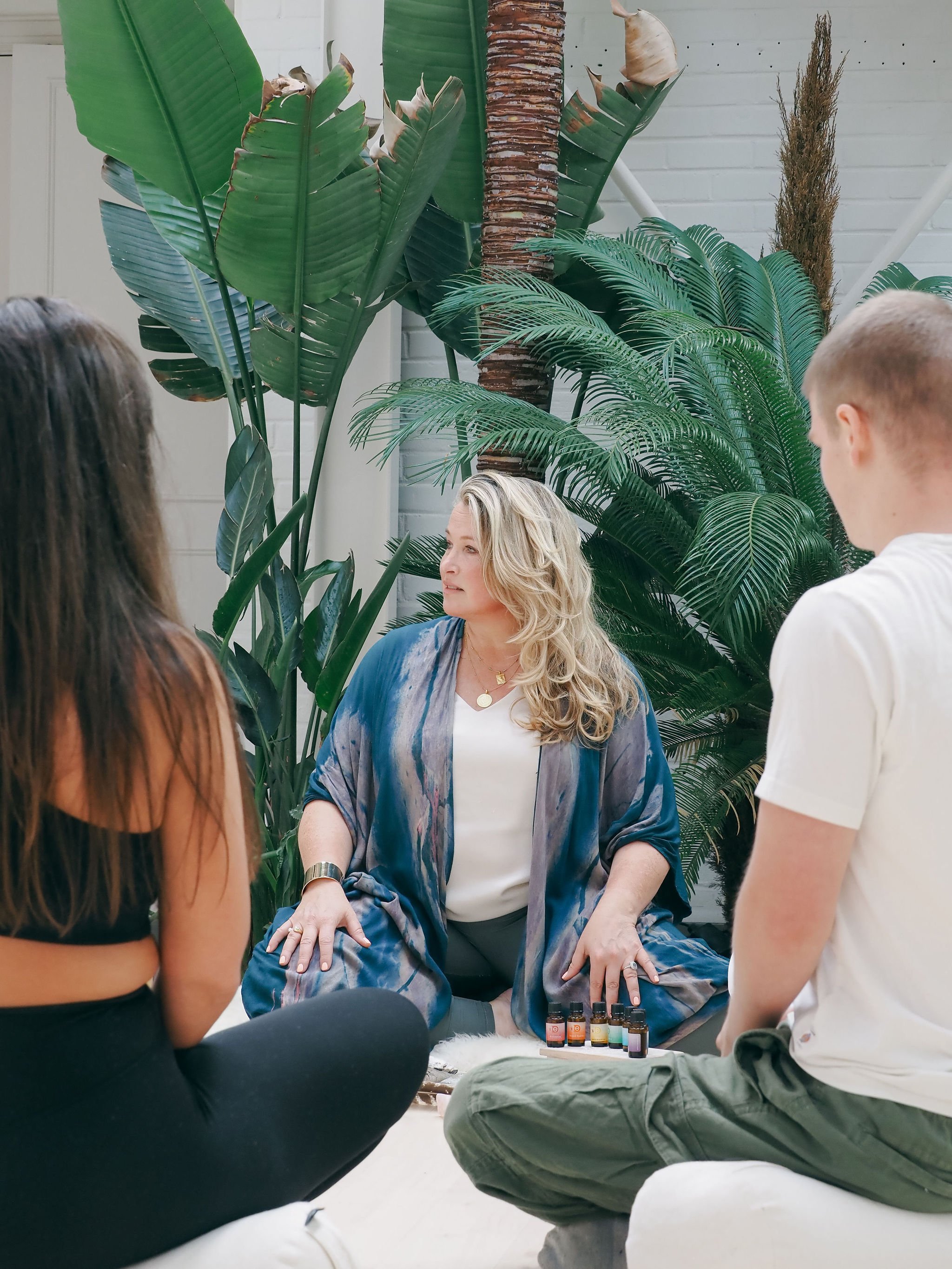 A woman seated, surrounded by three standing people, in front of large green tropical plants. The woman appears to be conducting a demonstration with small bottles of essential oils placed on a table before her.