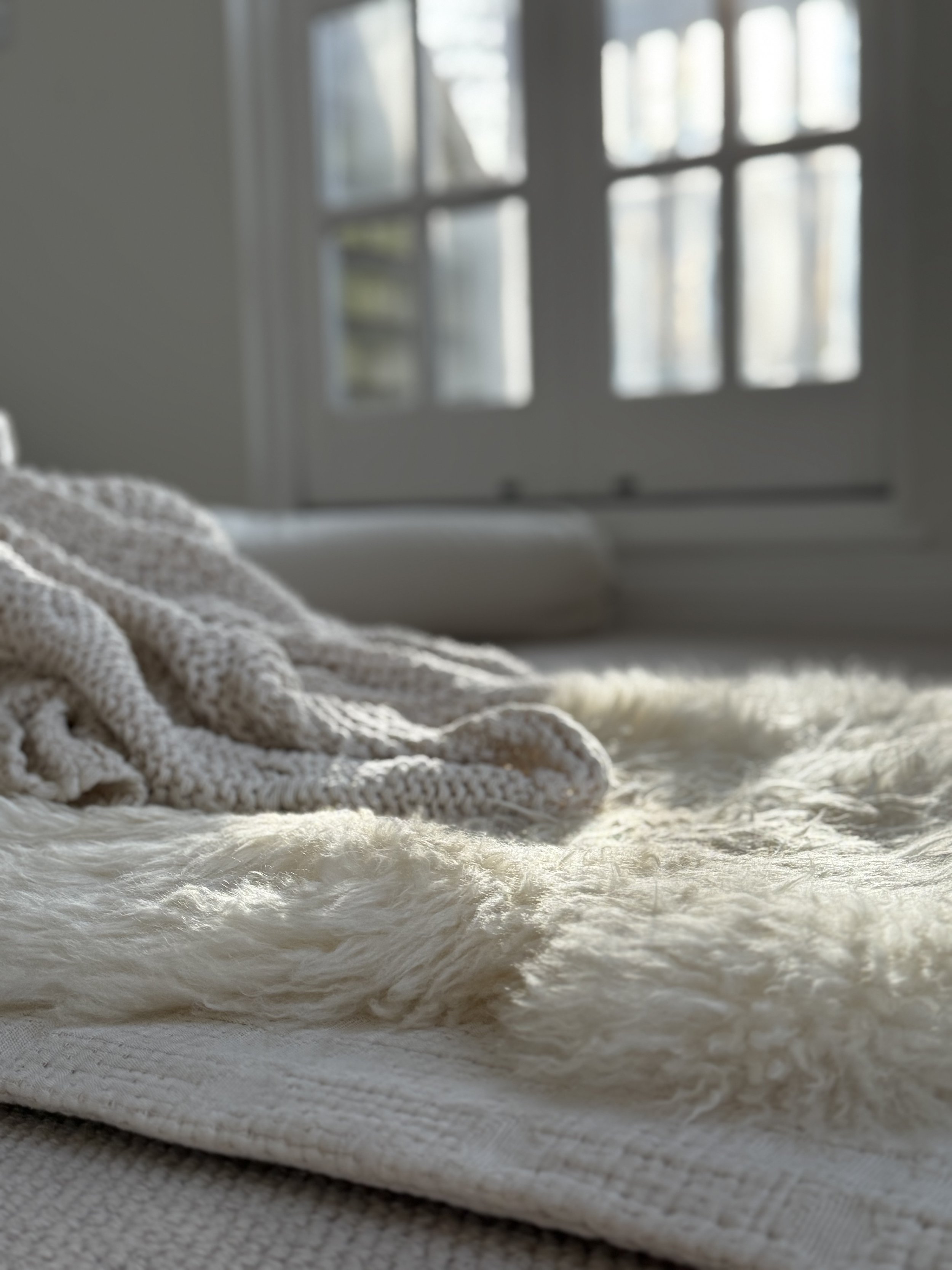 Close-up of a textured, beige fabric blanket on a bed with a blurred window in the background.