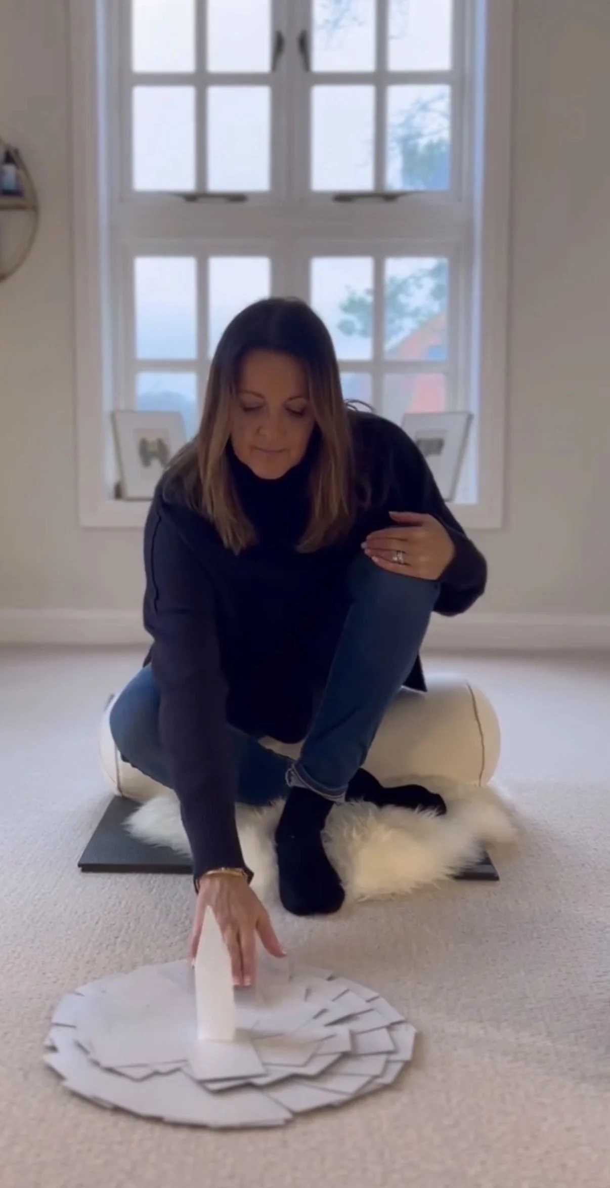 Woman practicing yoga in a room with large window, kneeling on a furry rug, surrounded by papers, with her hand reaching towards the papers.