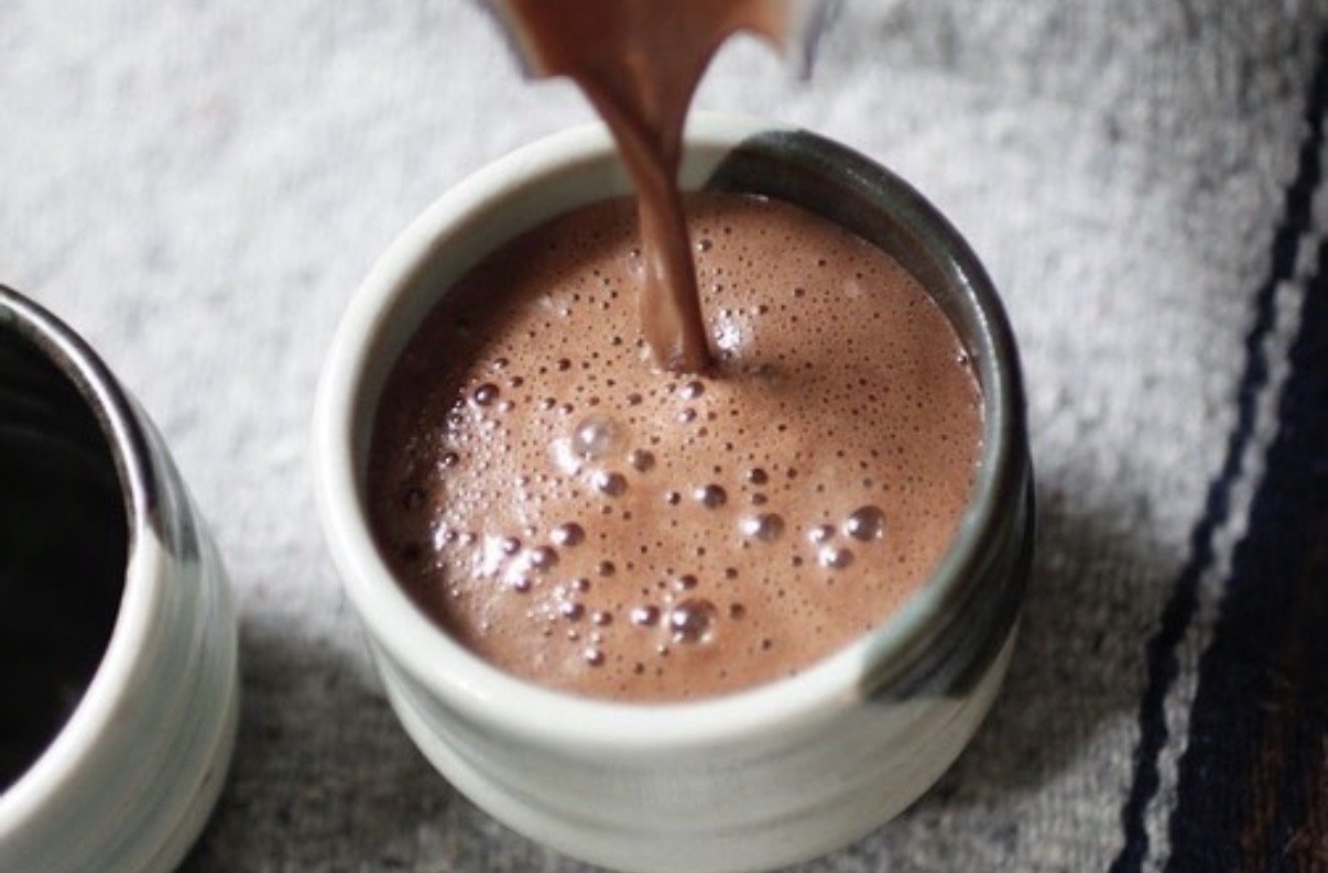 Hot chocolate being poured into a white mug on a gray textured surface.
