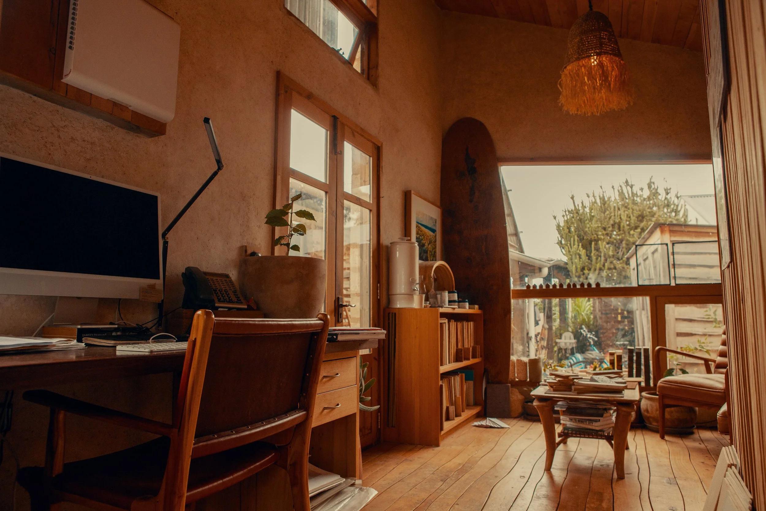 A cozy indoor space with wooden furniture, bookshelves, natural light coming through large windows, and a view of a backyard with trees and outdoor furniture.