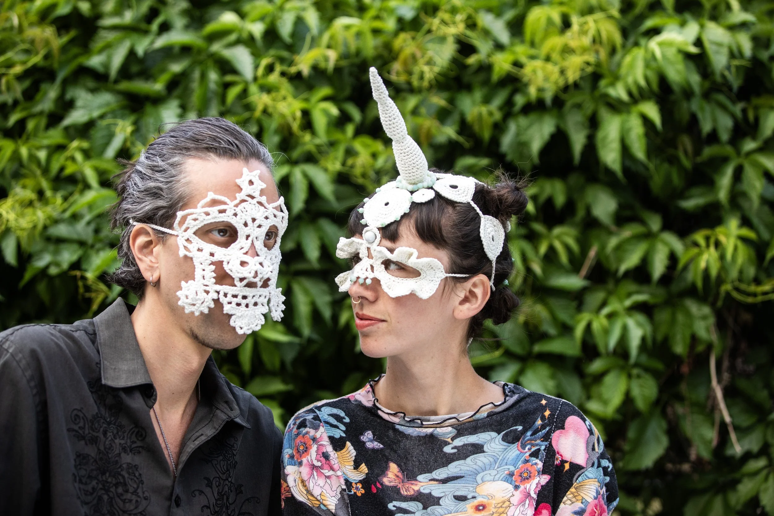 Two people wearing one of a kind crochet masks. One is a full skull face mask and the other is a unicorn headpiece.