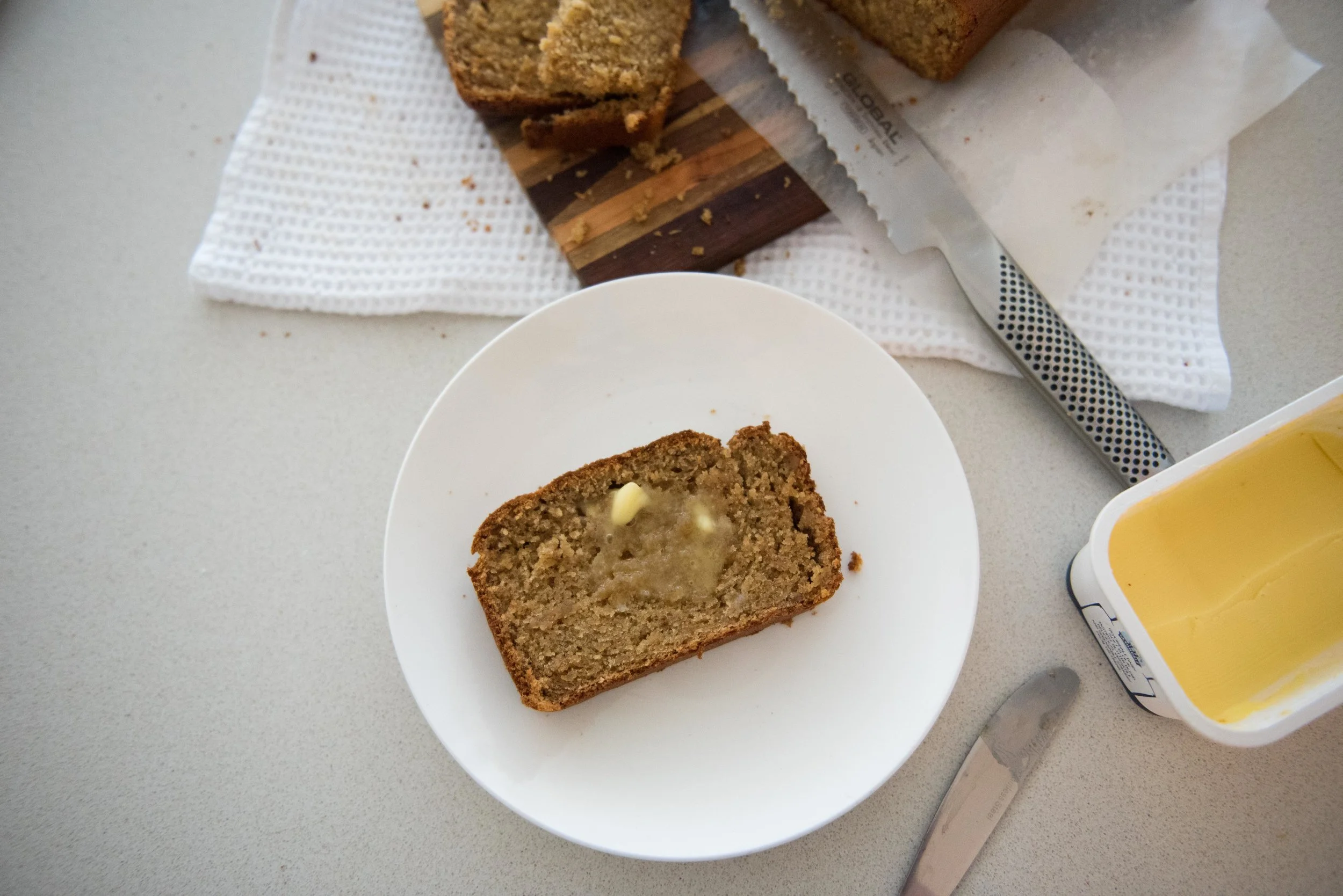 Banana Bread with context. You can see it has been freshly cut and butter has been melted through. I love the realness of this shot, it's not set up to the nth degree. There's crumbs, store bought butter and a buttery knife. Looks scrumptious!