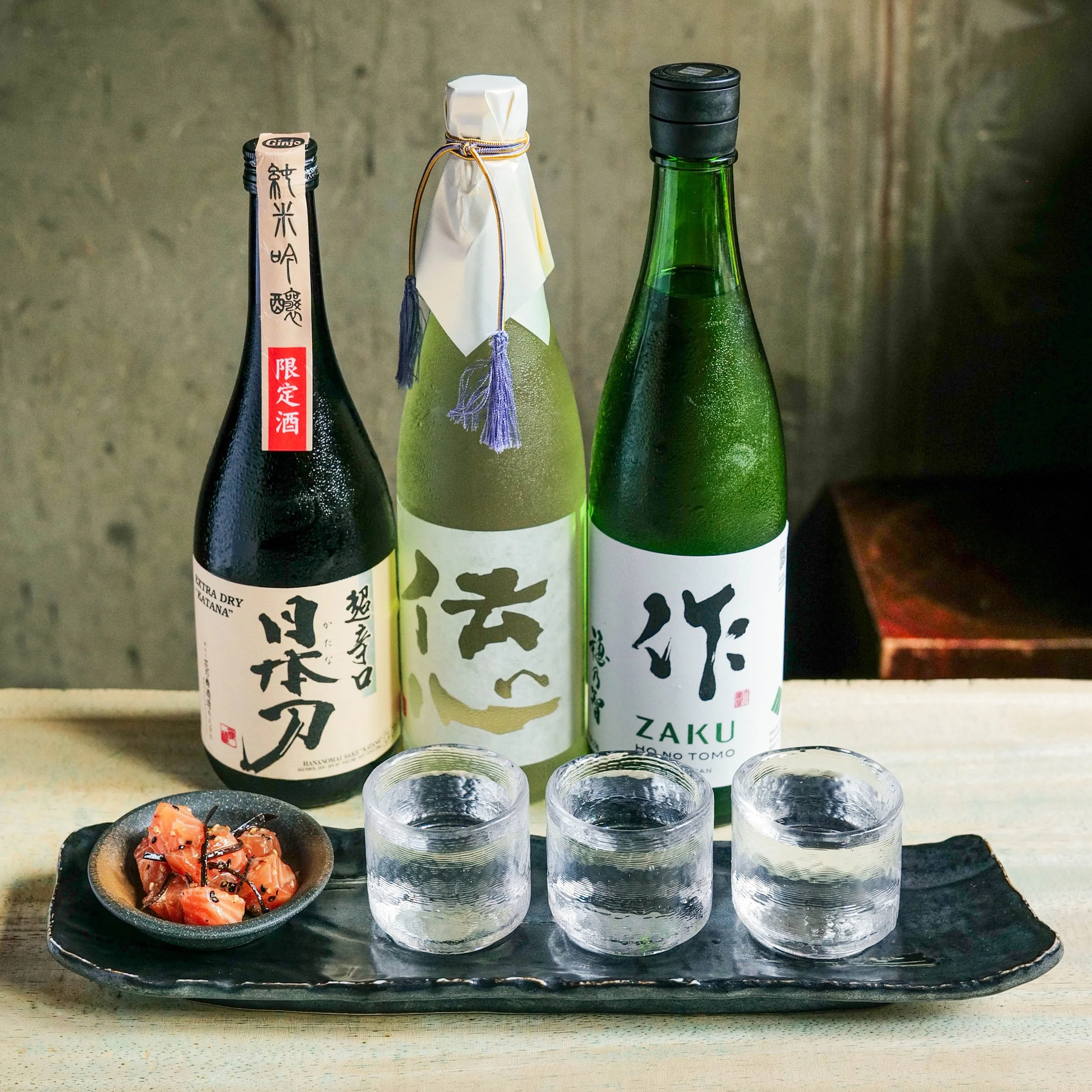 Three bottles of sake on a black rectangular tray, with a small dish of sliced salmon sashimi drizzled with sauce and sprinkled with black sesame seeds in front.