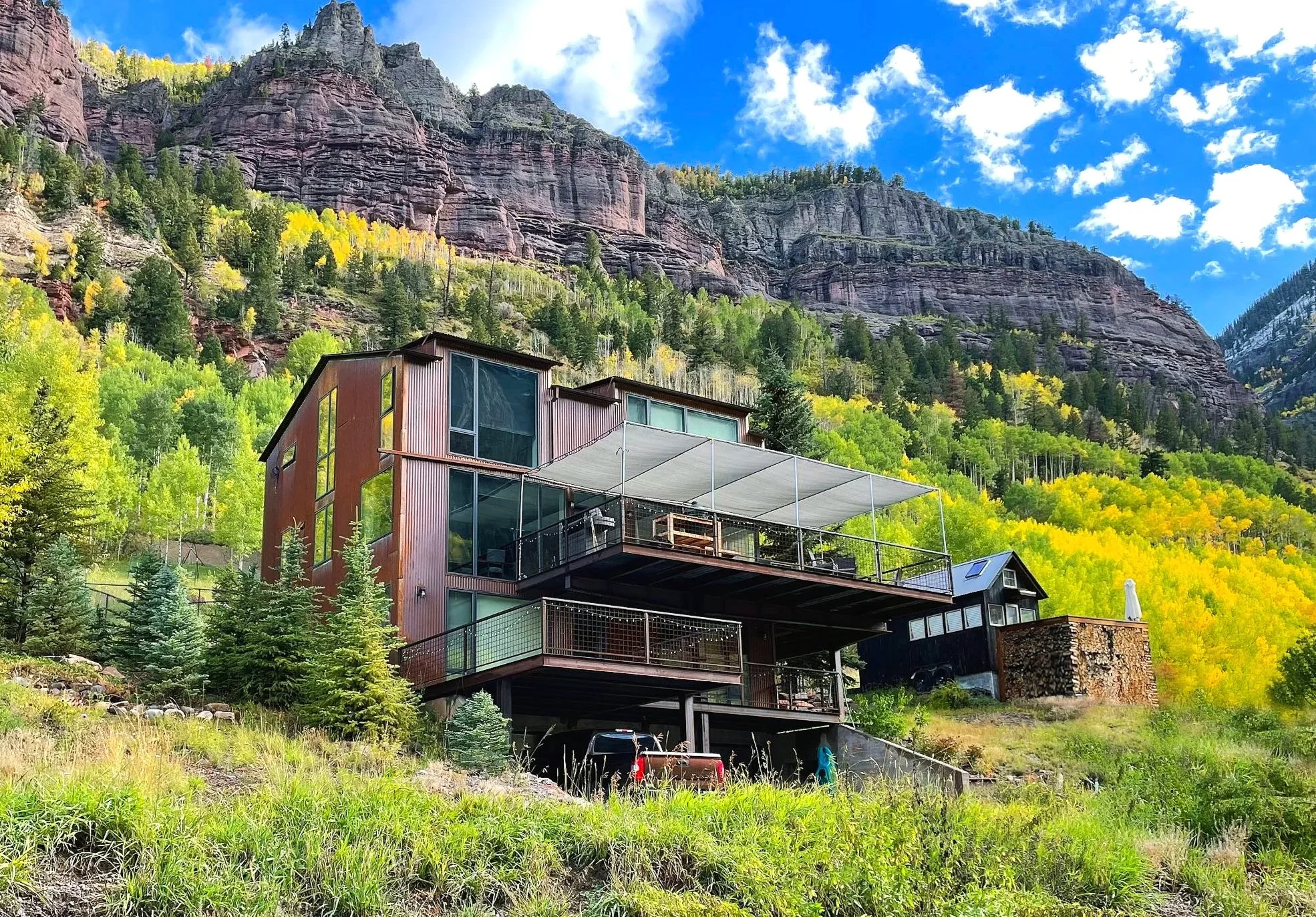 Modern multi-story house with large glass windows and a balcony, situated on a hillside with a forest of green and yellow trees at the base of a tall, rocky mountain under a partly cloudy blue sky.