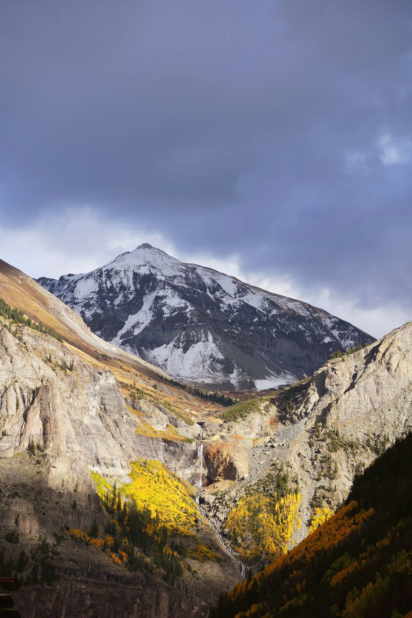 Snow-capped mountain in a valley with colorful autumn trees and rocky cliffs, cloudy sky overhead.