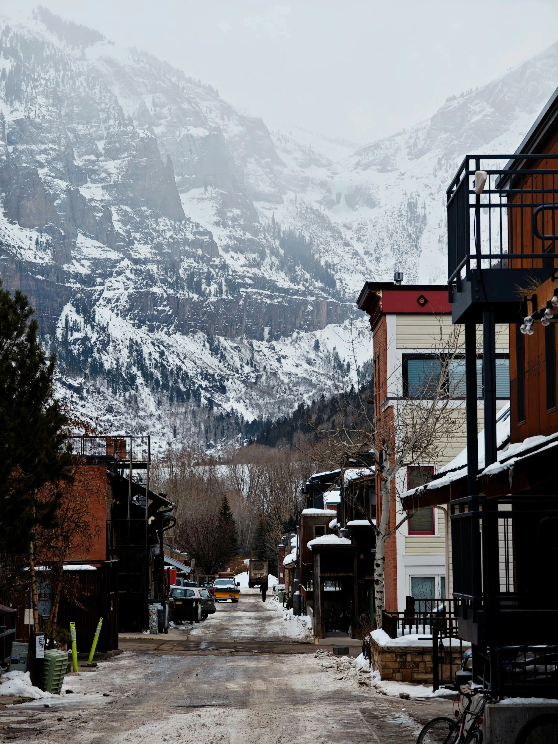 A snowy mountain town street with buildings on either side, parked cars, a person walking, snow on the ground, and mountains in the background.