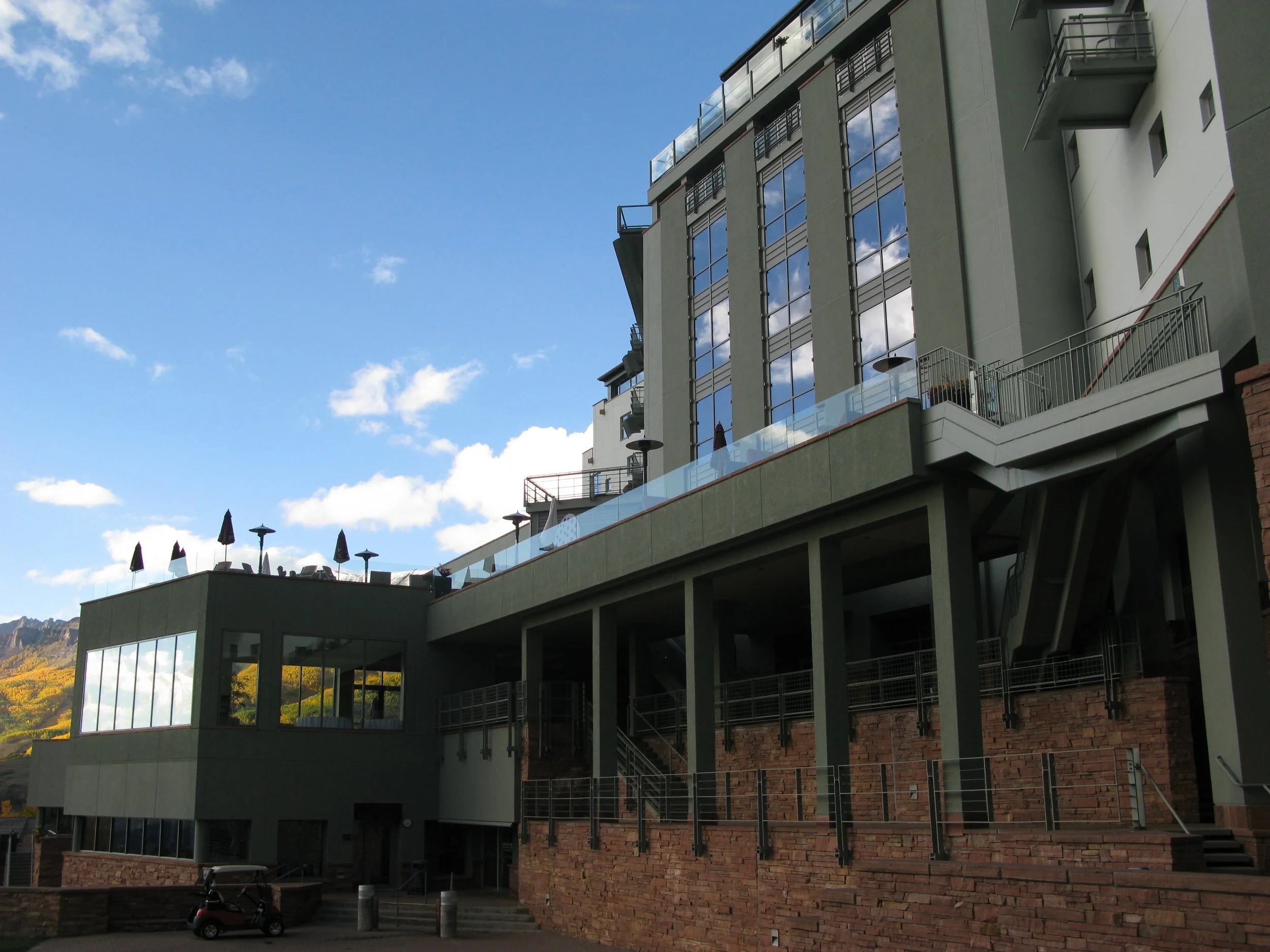 Multi-story modern building with large glass windows reflecting the sky and clouds, set against a blue sky with some clouds, and a scenic mountainous landscape in the background.