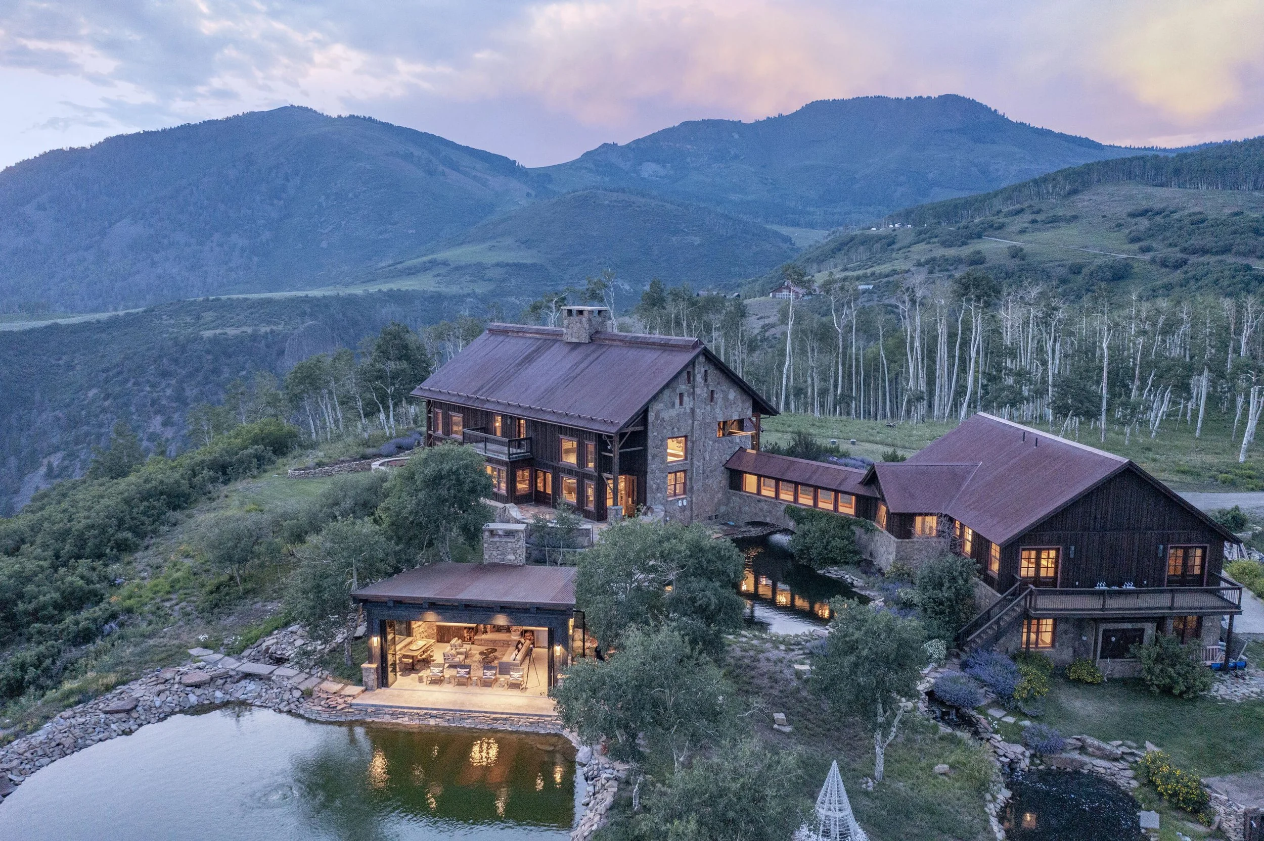 A large mountain landscape with a rustic house and a pond in the foreground, surrounded by trees and hills under a cloudy sky.