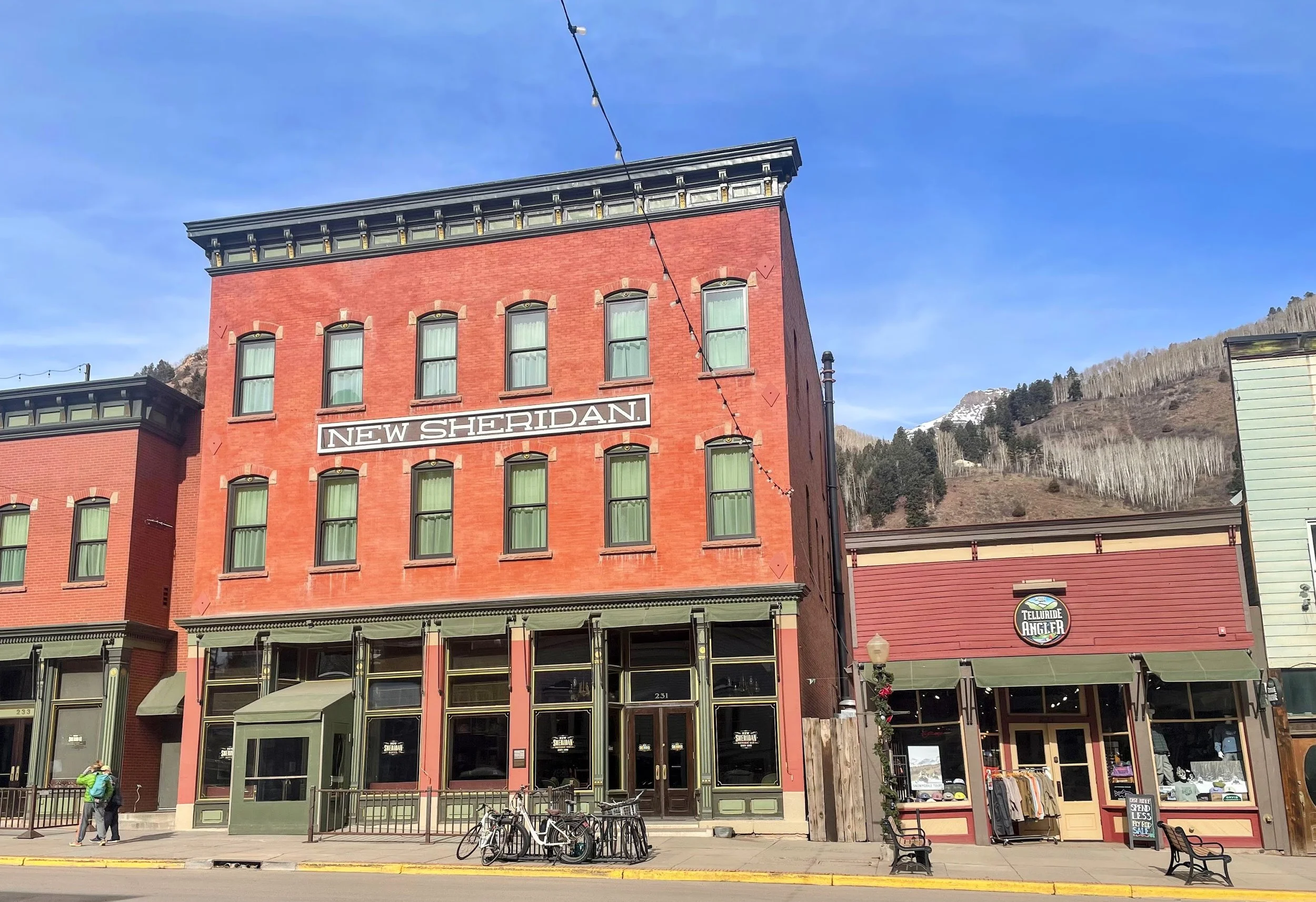 Red brick building with a sign that says "New Sheridan" and green window curtains. There are bicycles parked in front, and a person with a green jacket and backpack walking nearby. The building has upper-story windows and a storefront on the ground level. The background features a mountain with some snow and trees.