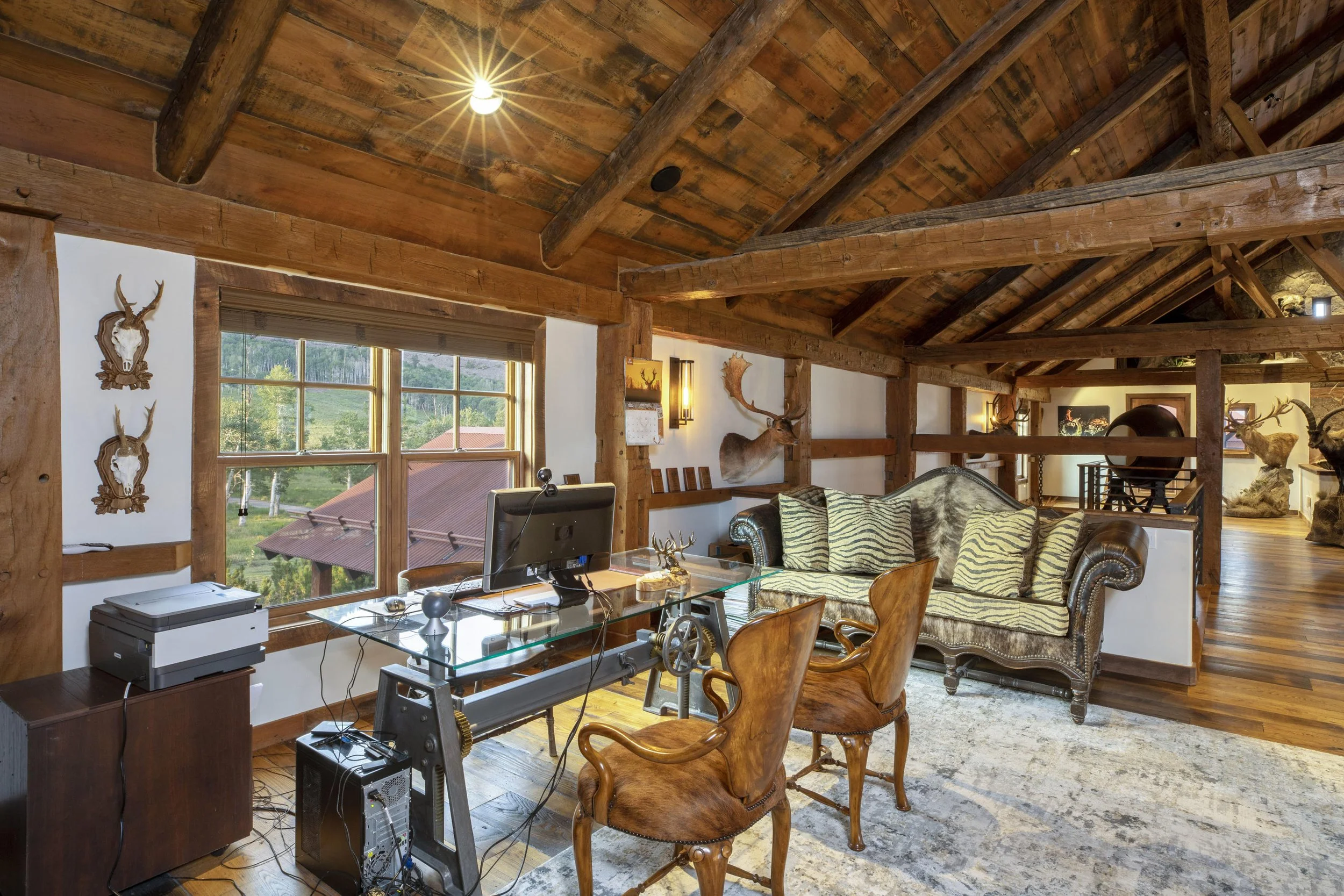 Interior view of a rustic log cabin living room with exposed wooden beams, decorated with mounted animal heads, including deer, and featuring large windows showing a green outdoor landscape.