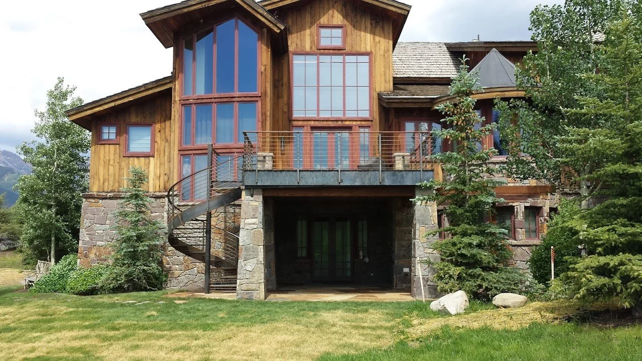 A large multi-story house with a wooden exterior, stone foundation, and large windows, surrounded by trees and a green lawn.