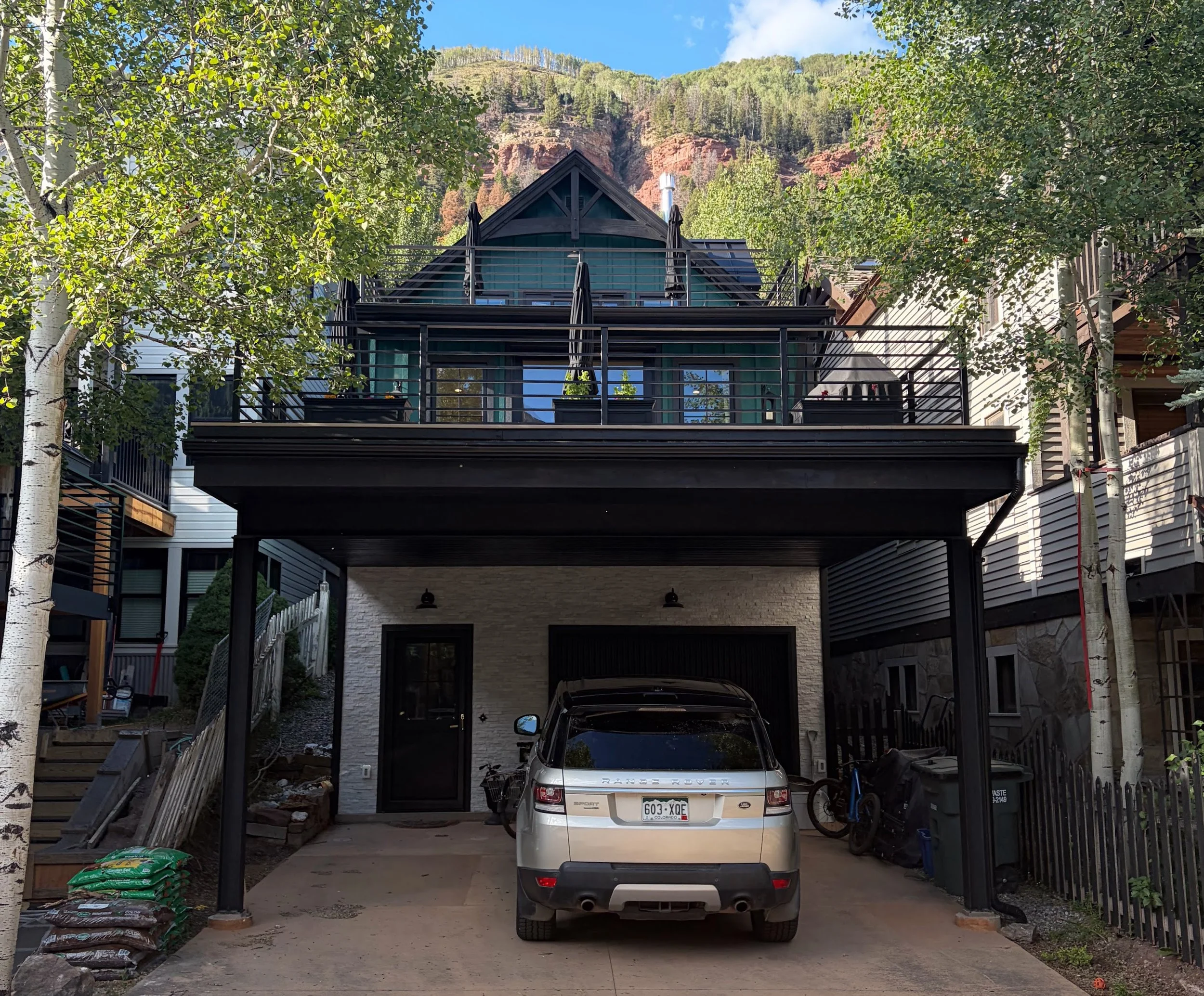 A house with multiple decks and a black metal railing, with trees and a mountain in the background. There is a silver Range Rover parked in the driveway.