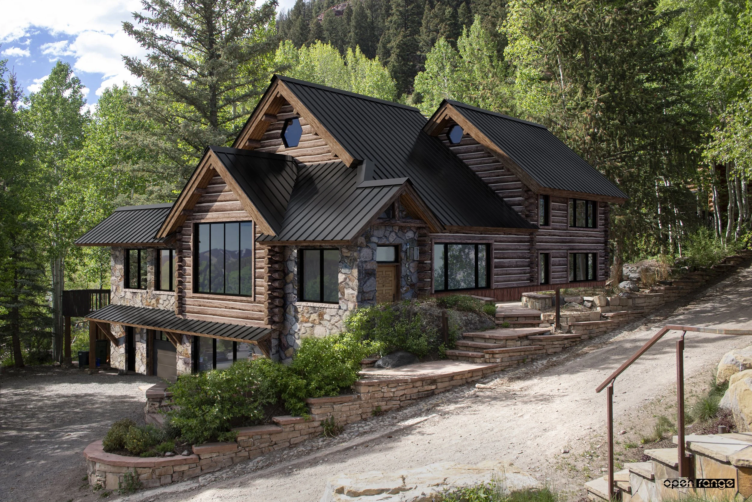 A rustic log cabin-style house with a black metal roof and large windows, set amidst tall green trees and a mountainous forest landscape.