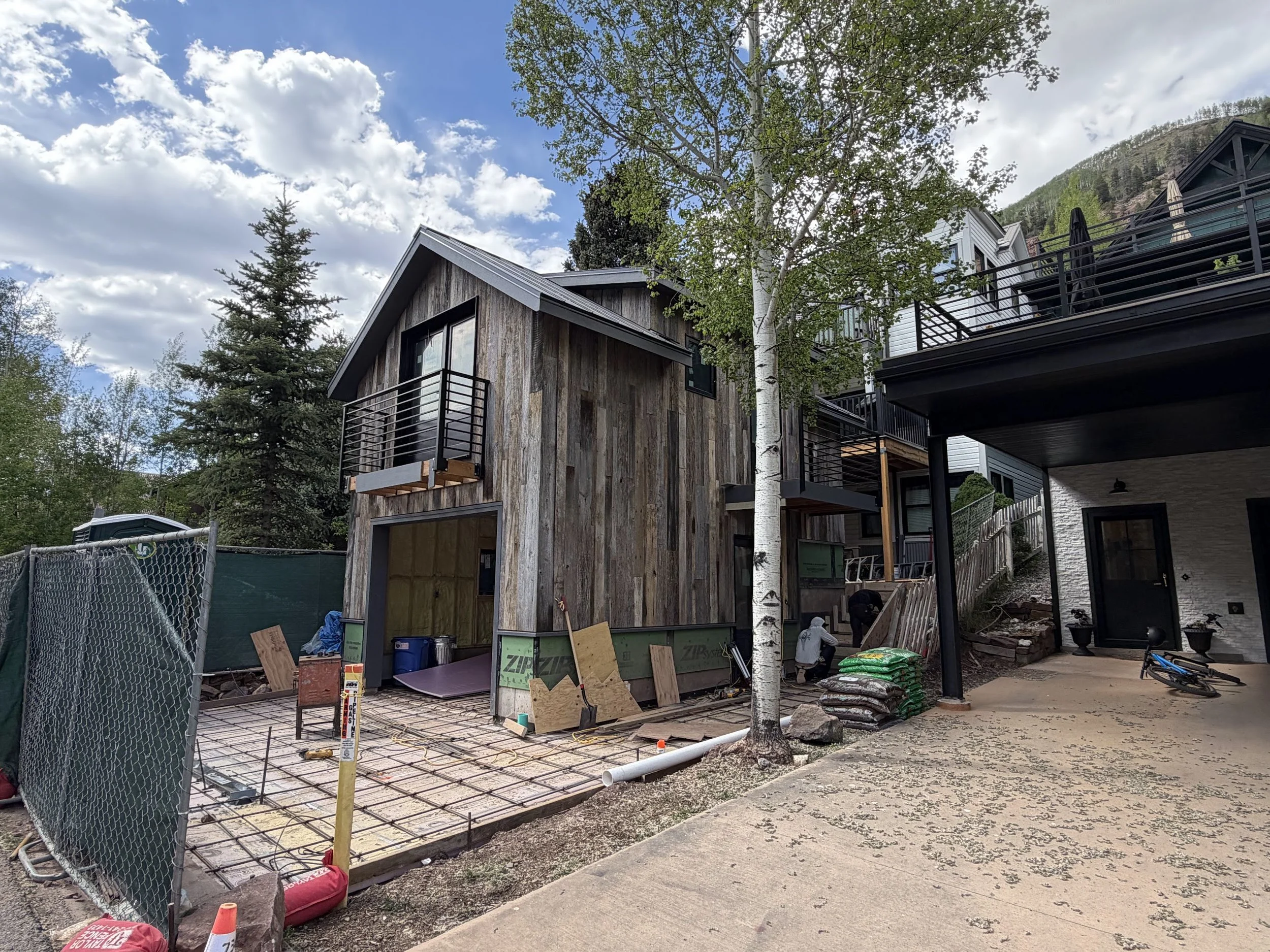 Under-construction residential building with wooden and white siding, surrounded by trees and construction supplies.