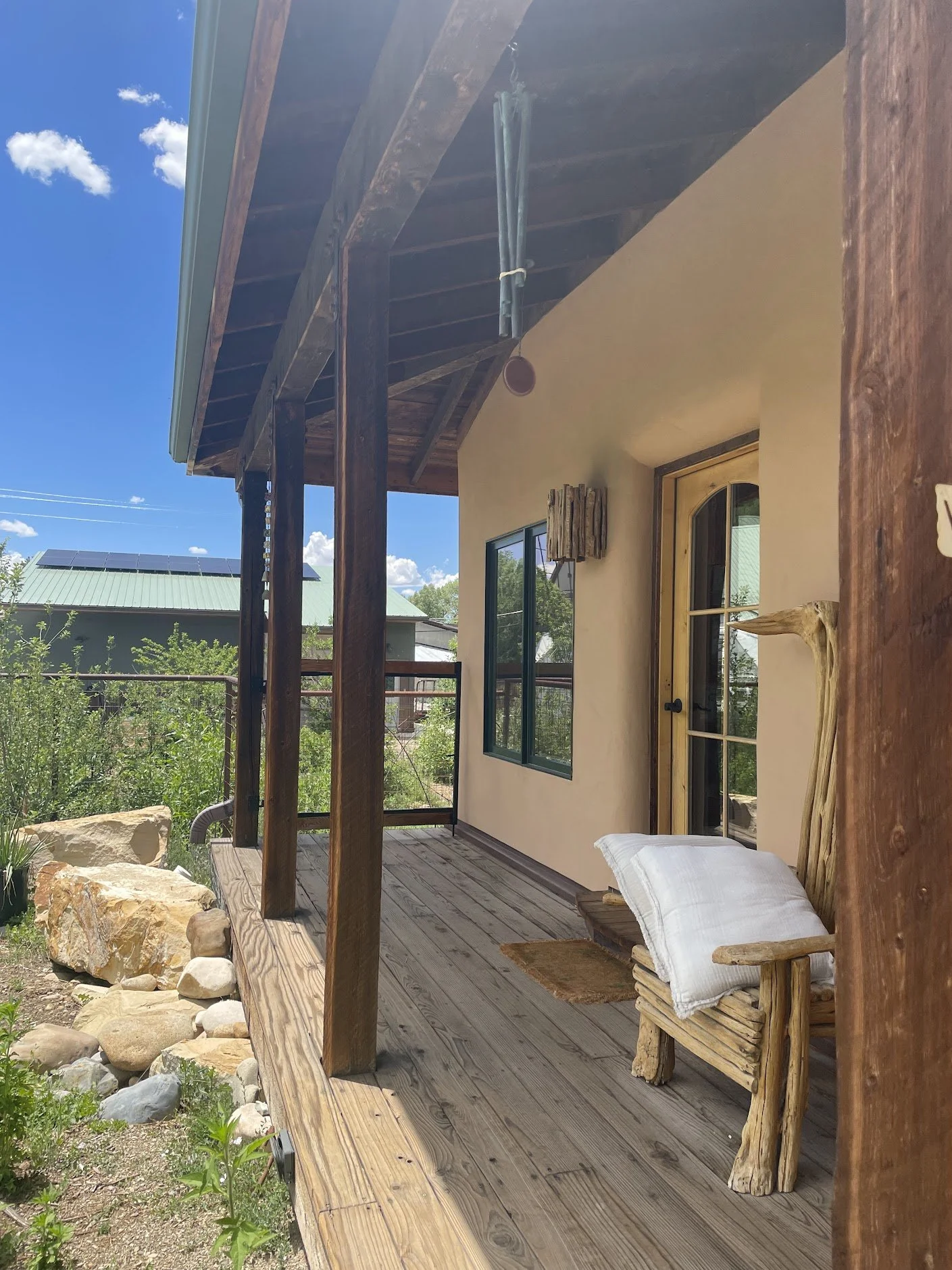 A cozy outdoor porch with wooden flooring and a railing, featuring a rustic wooden chair with a white pillow, adjacent to a door with glass panes. The porch is attached to a house with a stucco exterior, and there are rocks and greenery surrounding i