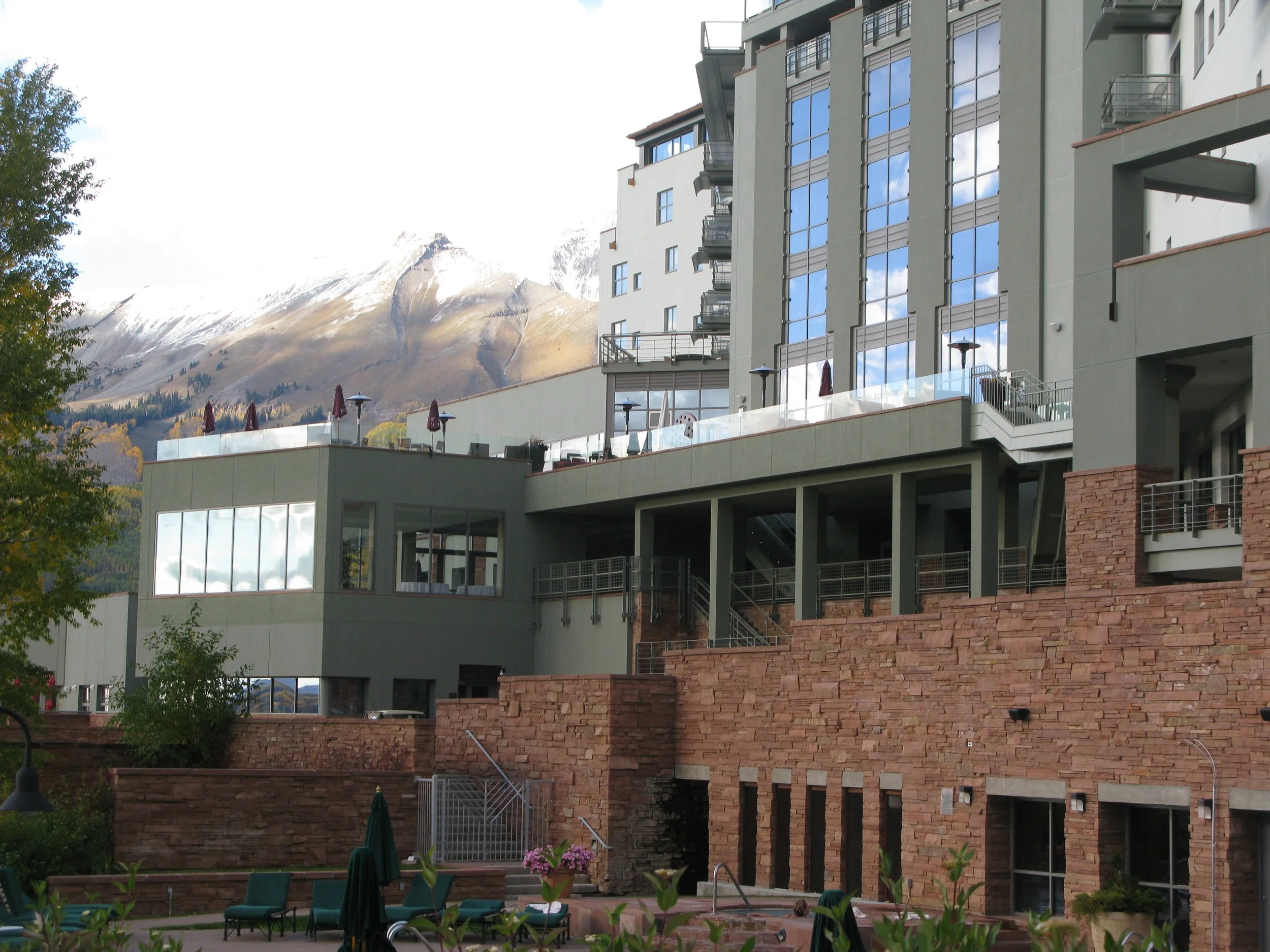 Modern multi-story residential building with large reflective windows, balconies, and outdoor patio areas, set against a mountain landscape with snow-capped peaks in the background.