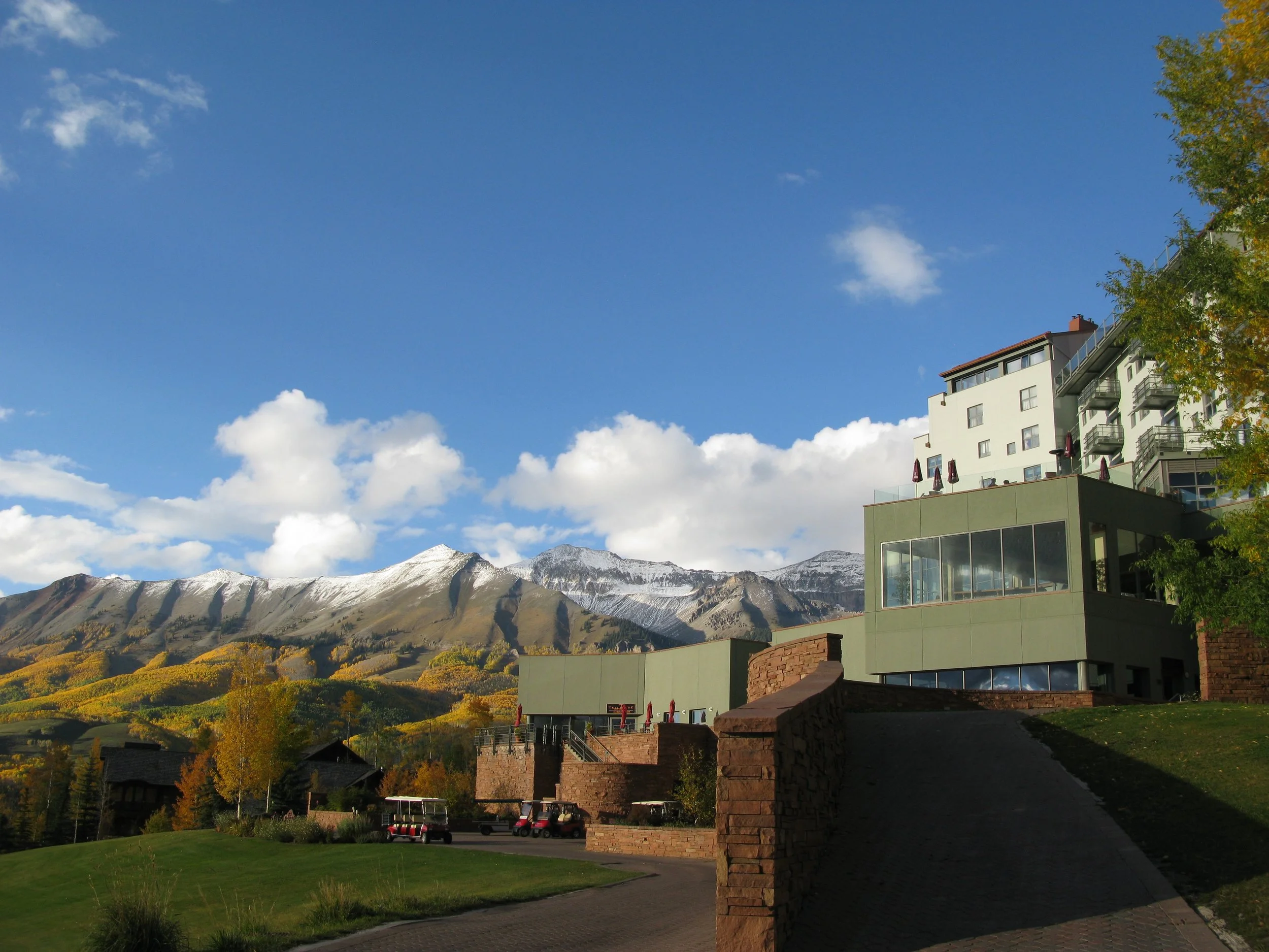 A large modern green building with multiple levels and glass windows is situated on a hillside, with autumn-colored trees surrounding it. In the background, snow-capped mountains are visible under a blue sky with scattered clouds.