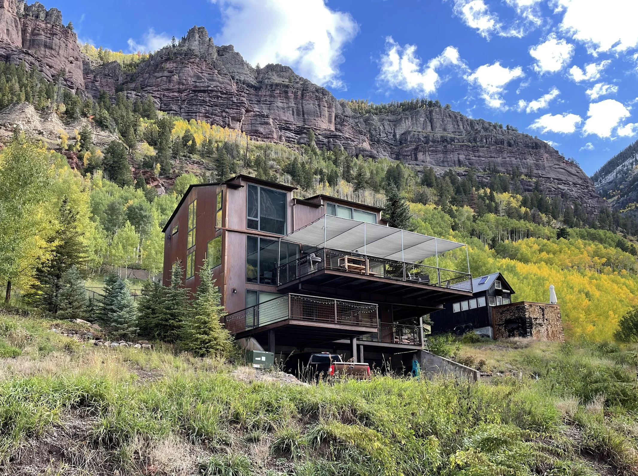 Modern multi-story house with large windows and outdoor deck situated on a hillside at the base of a rugged mountain with forest and rocky cliffs, under a bright blue sky with scattered clouds.