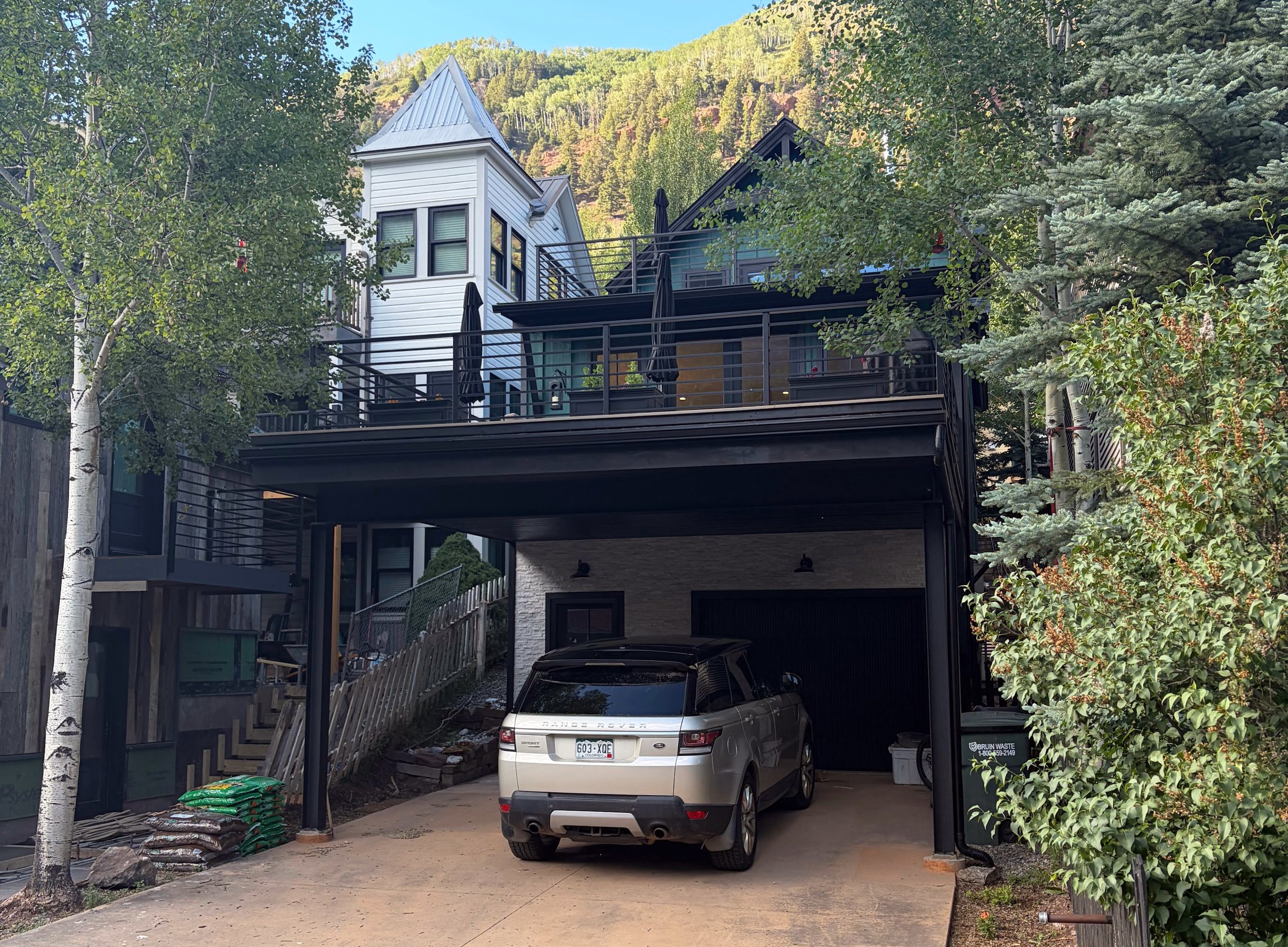 A multi-story house with a balcony, metallic roof, and large windows surrounded by trees and mountains in the background. There is a driveway with a white Range Rover parked underneath the house, and construction materials and a fence visible on the 