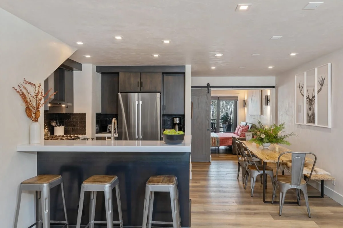 Open-concept kitchen and dining area with gray cabinetry, stainless steel refrigerator, wooden dining table with metal chairs, and decorated with plants and artwork.