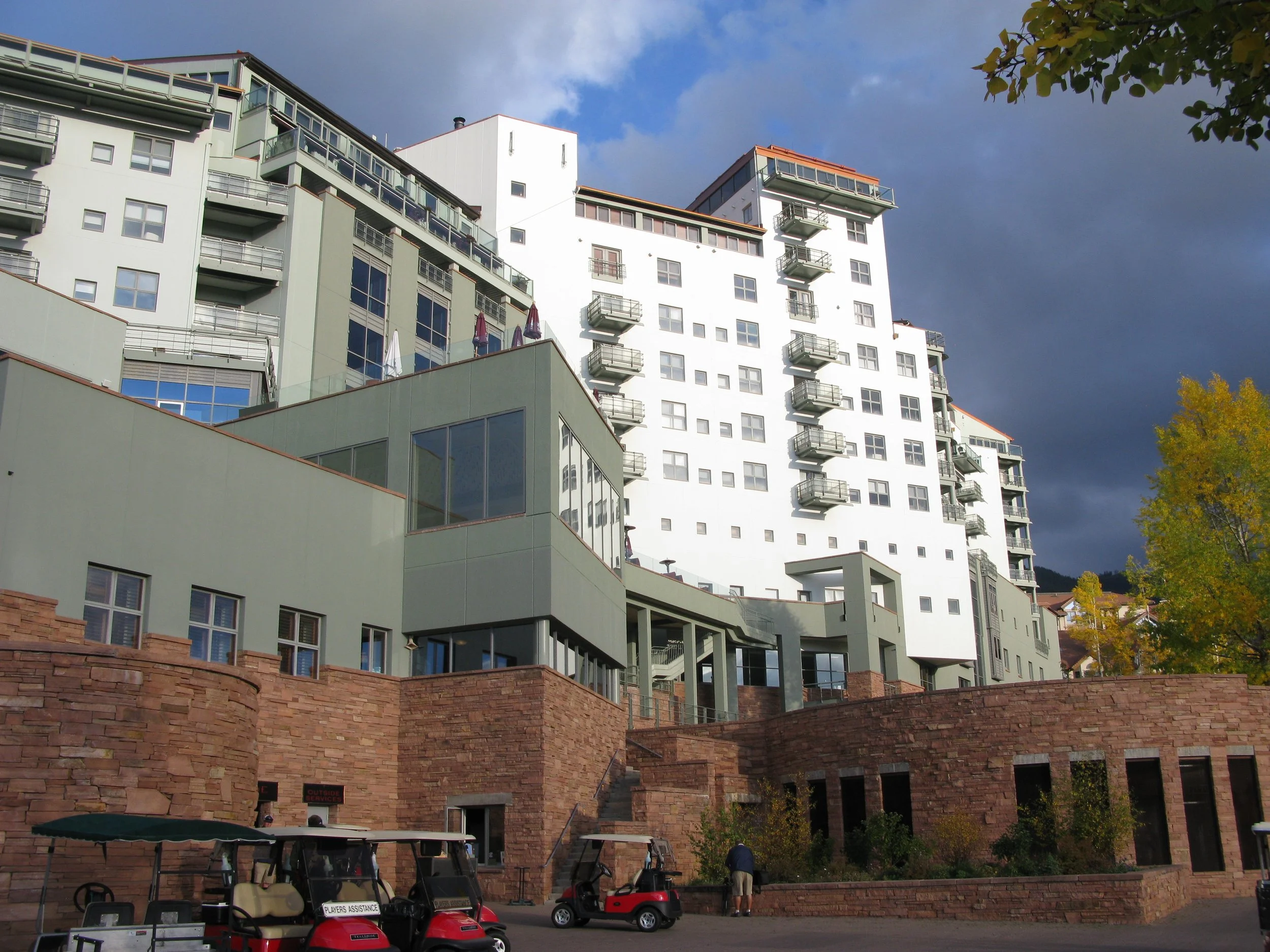 A modern multi-story residential building with balconies and large windows, set against a partly cloudy sky, with trees and golf carts in the foreground.
