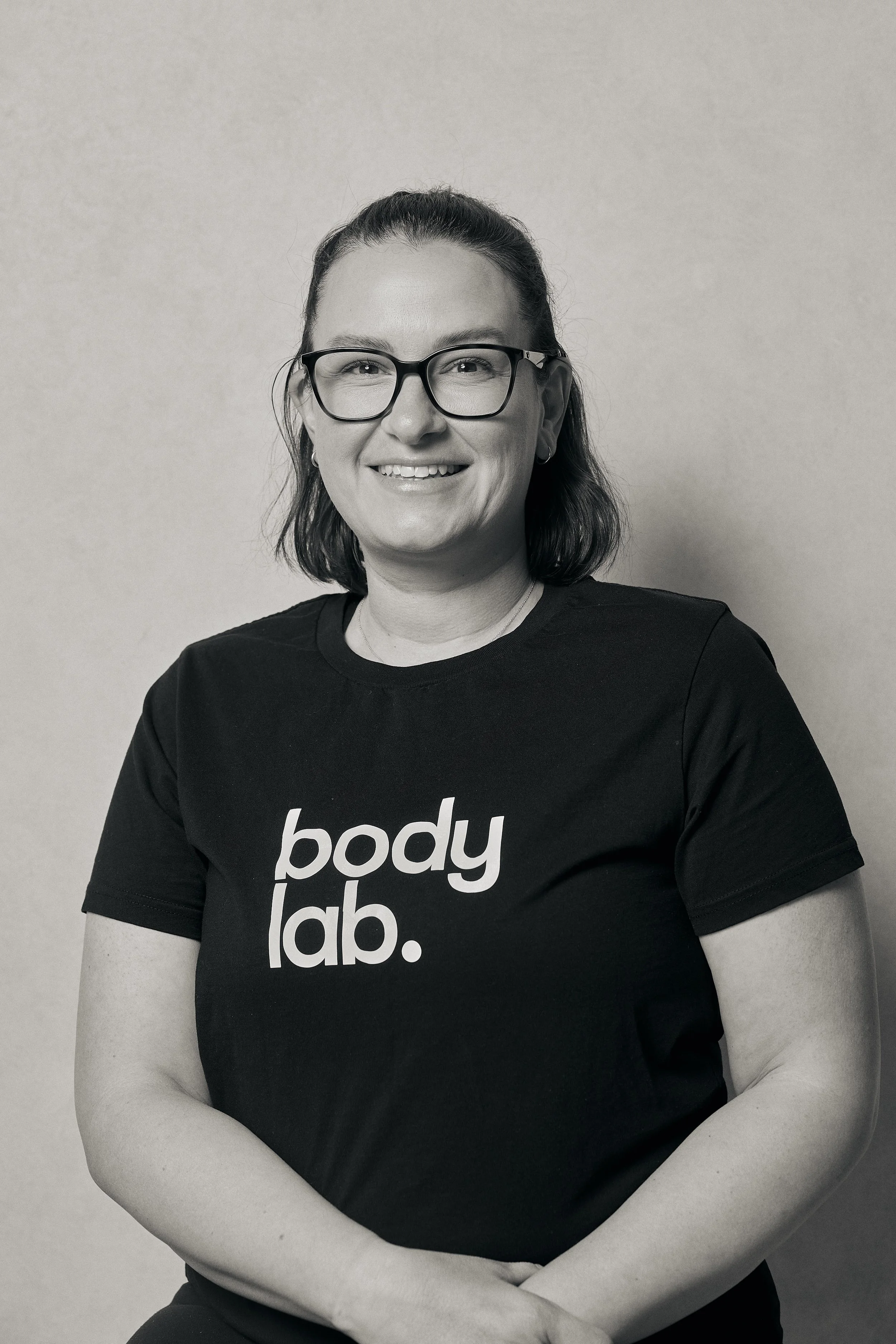 A woman with glasses, smiling, wearing a black t-shirt with 'body lab.'