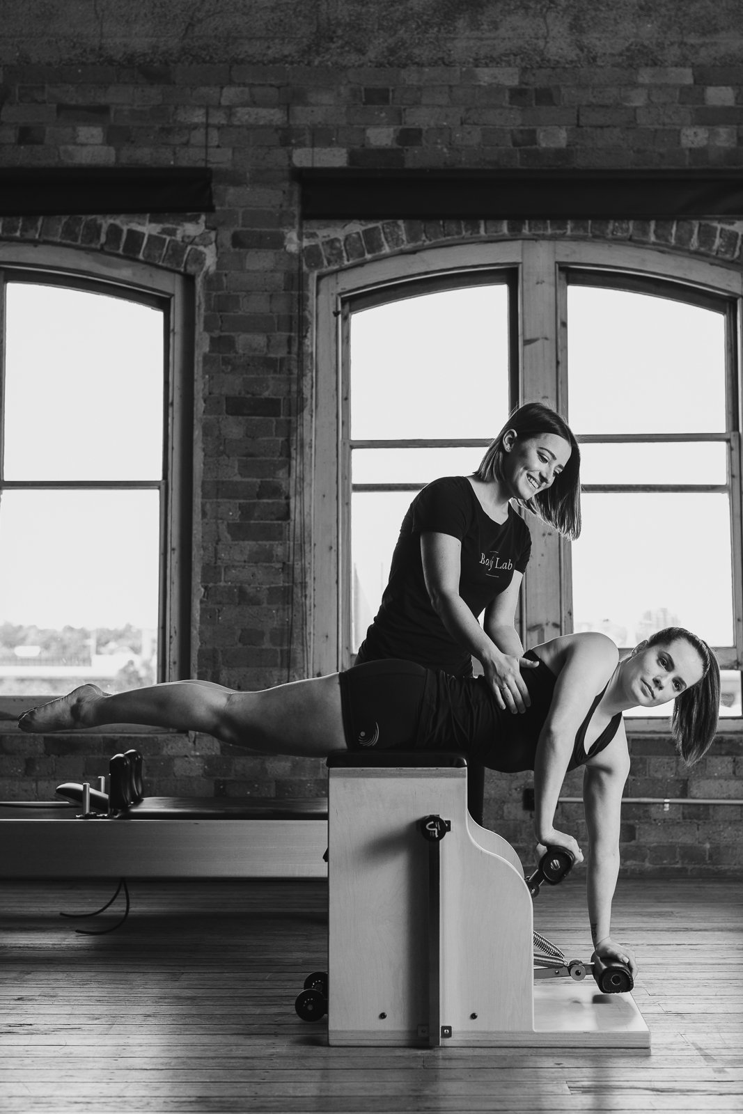 A woman performs a Pilates exercise on a reformer machine while a trainer assists her in a gym with large windows and brick walls.