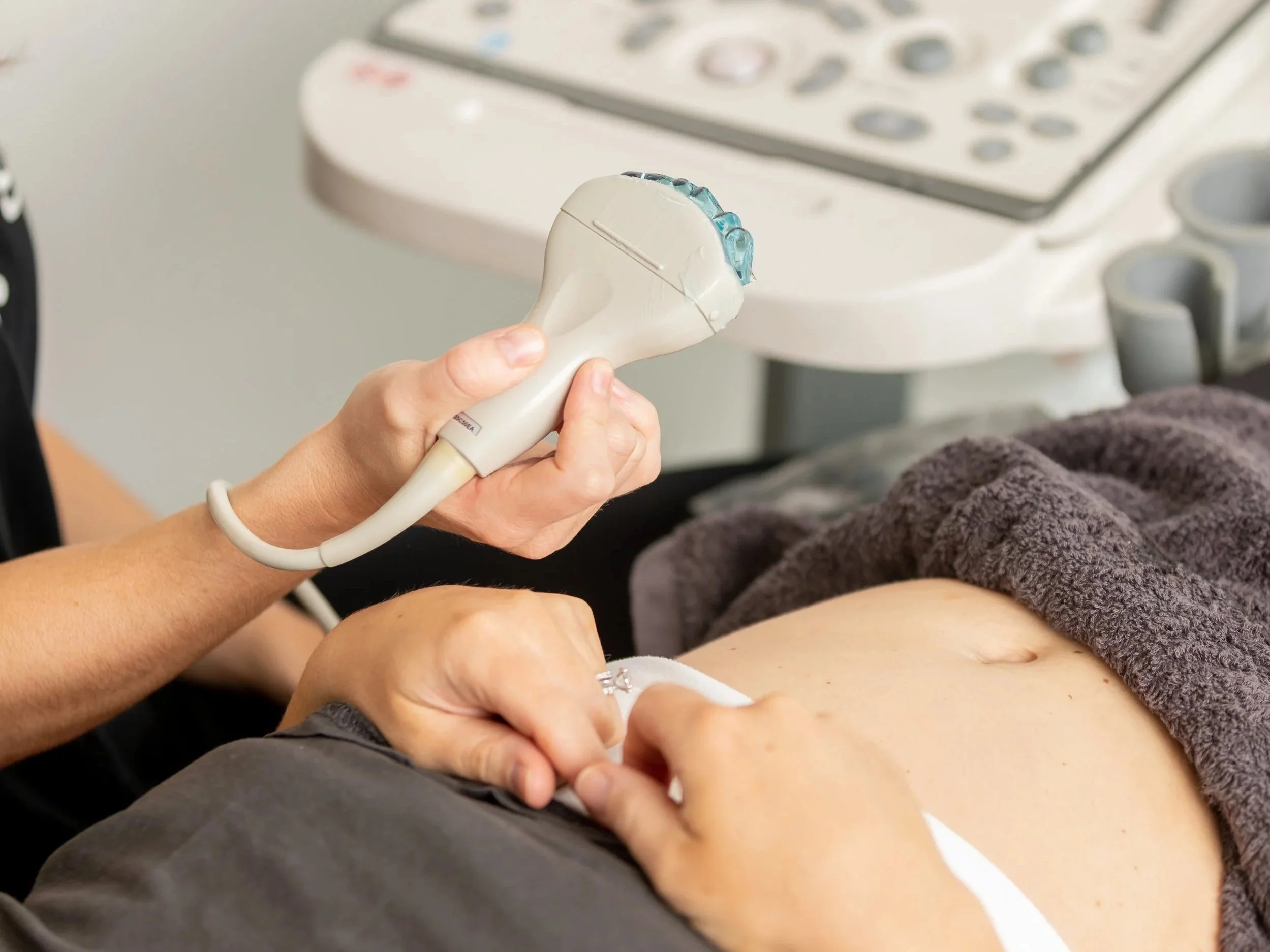 A healthcare professional performs an ultrasound on a patient's abdomen using a handheld probe, with an ultrasound machine in the background.