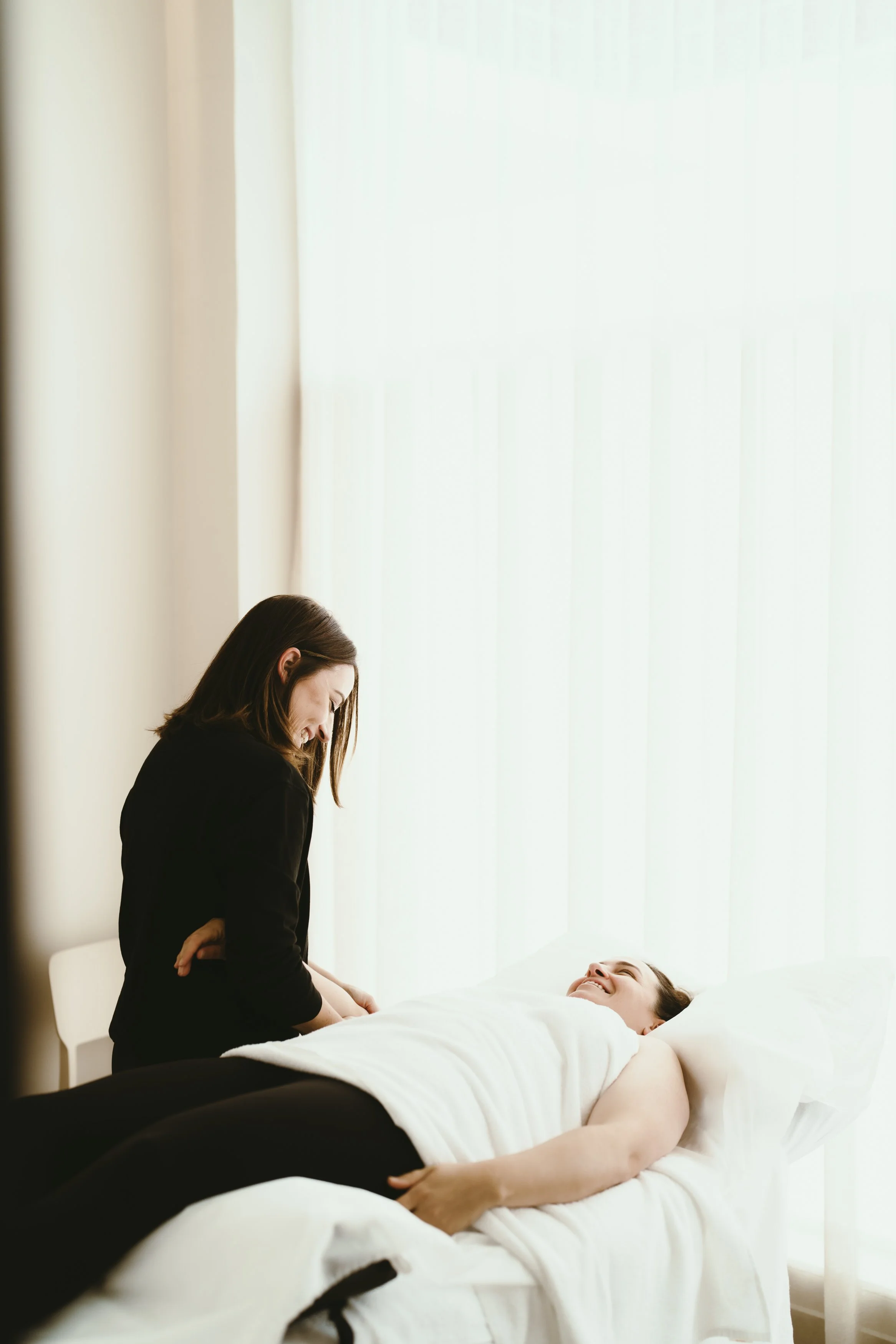 A physiotherapist leaning over a patient, both smiling and engaging in conversation in a brightly lit clinic room.