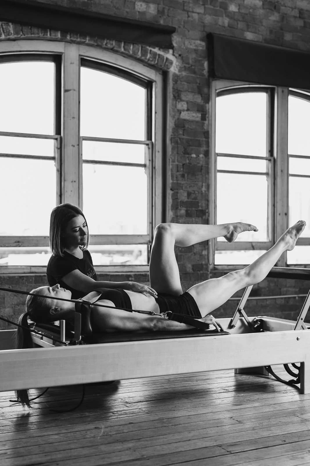 A woman performs a Pilates exercise on a reformer machine while an instructor assists her in a studio with brick walls and large windows.