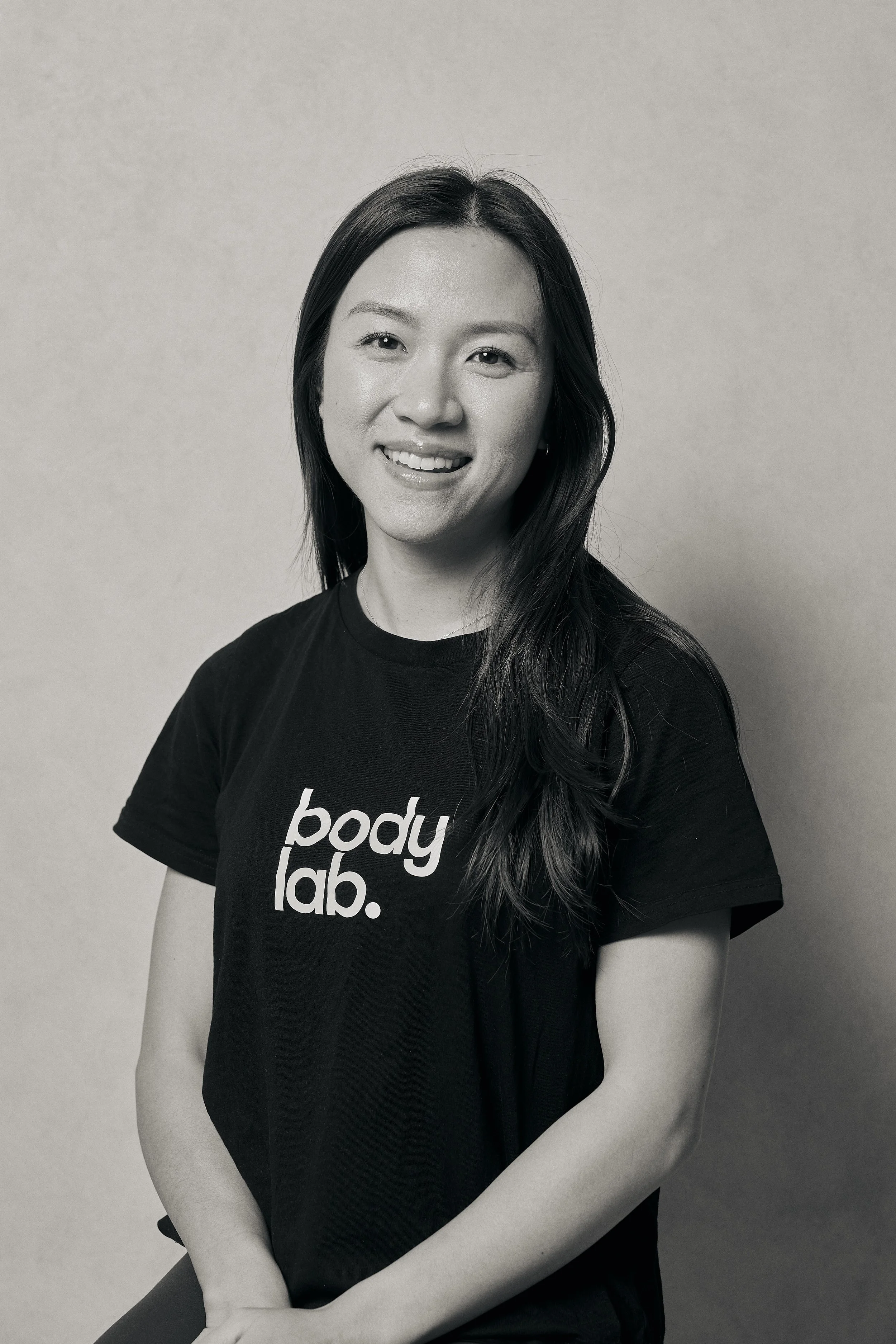 A smiling woman with long dark hair wearing a black t-shirt with 'body lab.' written on it, posing against a plain background.