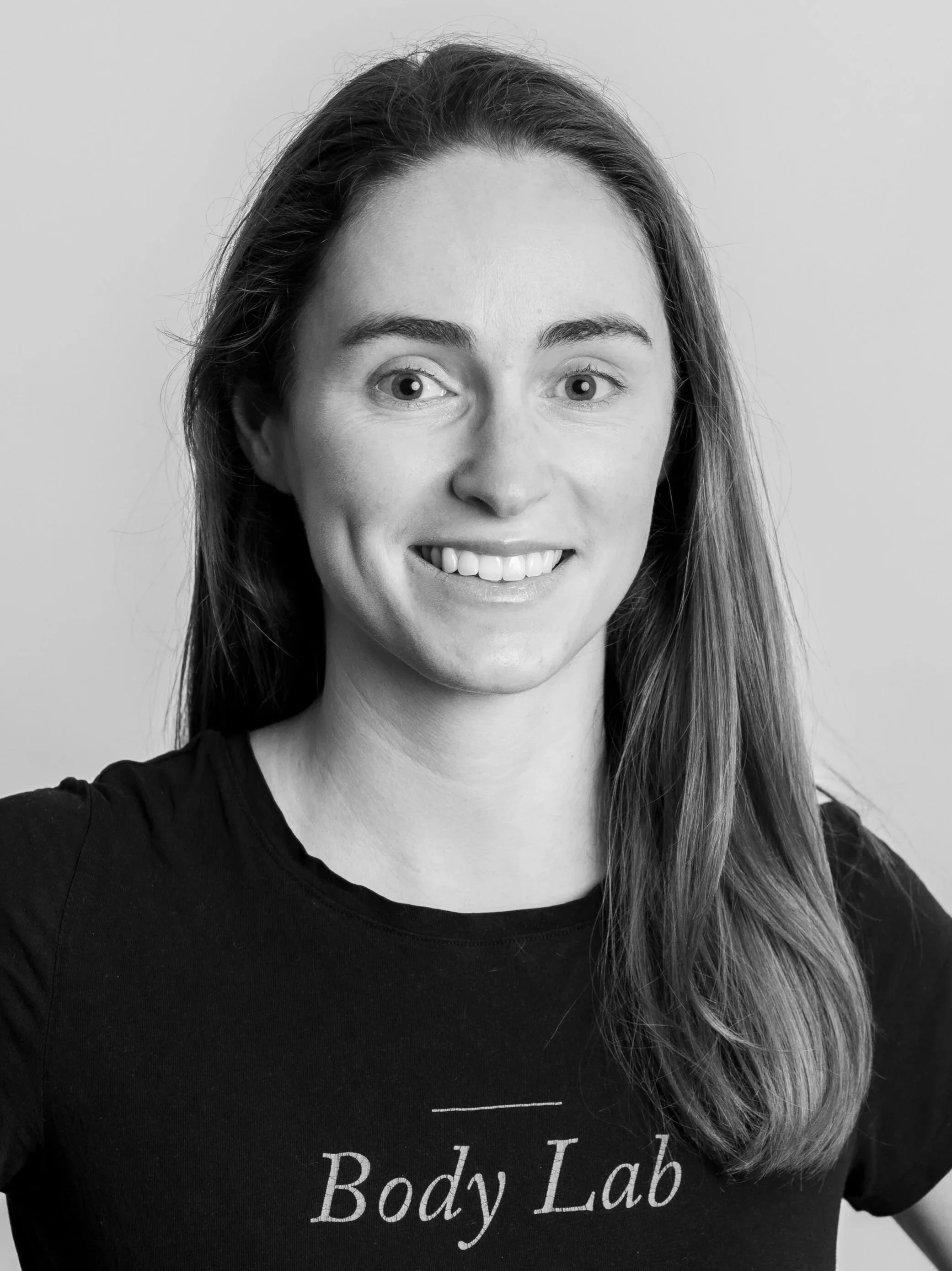 A woman with long hair smiling at the camera, wearing a T-shirt with "Body Lab" written on it, black and white photo.
