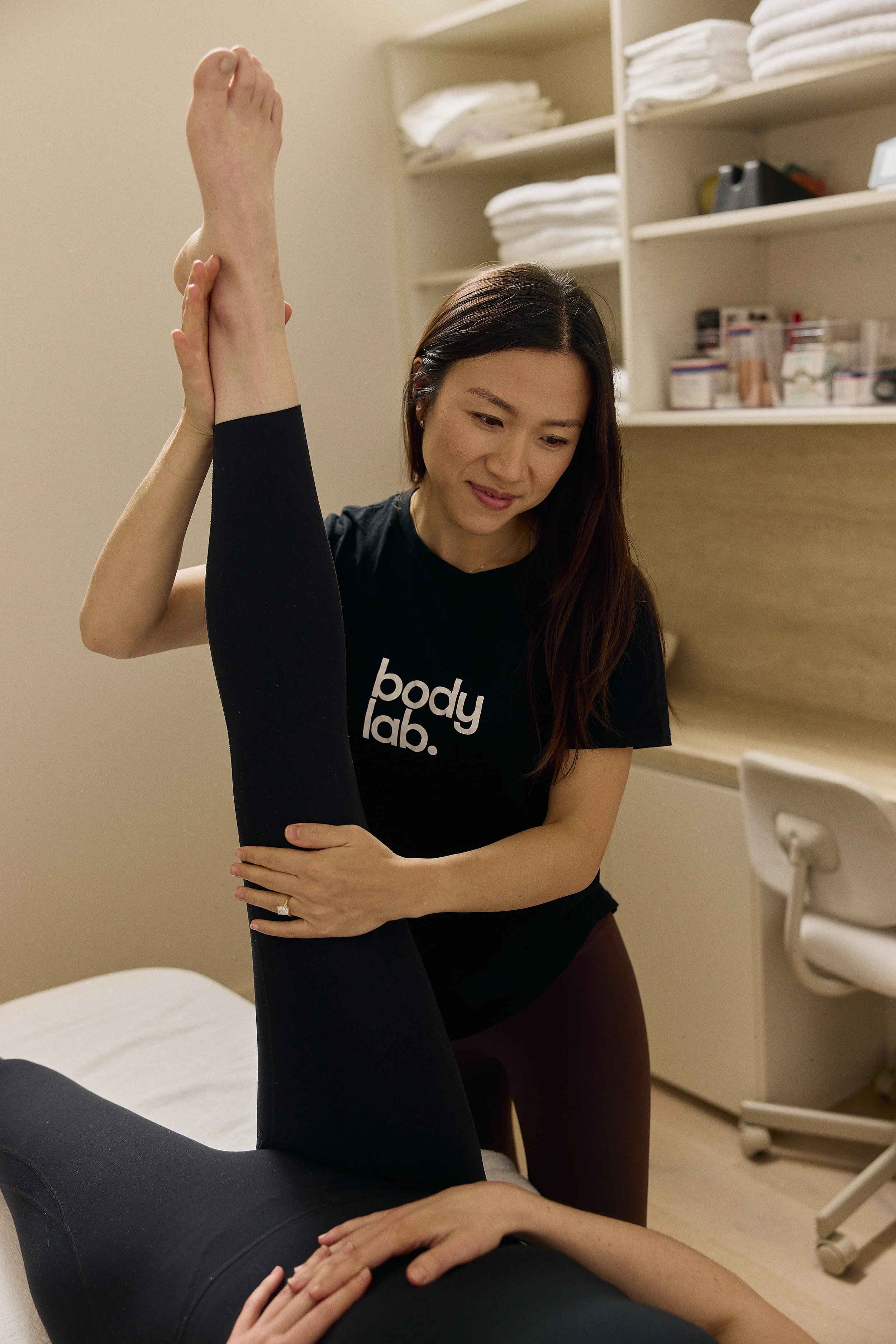 A woman in a black t-shirt with 'Body Lab' written on it,  stretching a patient's leg in a clinic or therapy room.
