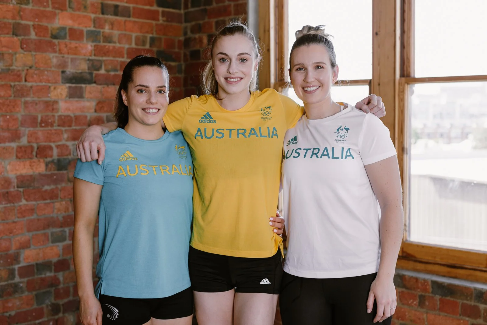 Three female athletes wearing Australia team shirts, smiling and standing arm-in-arm inside a brick-walled room with large windows.