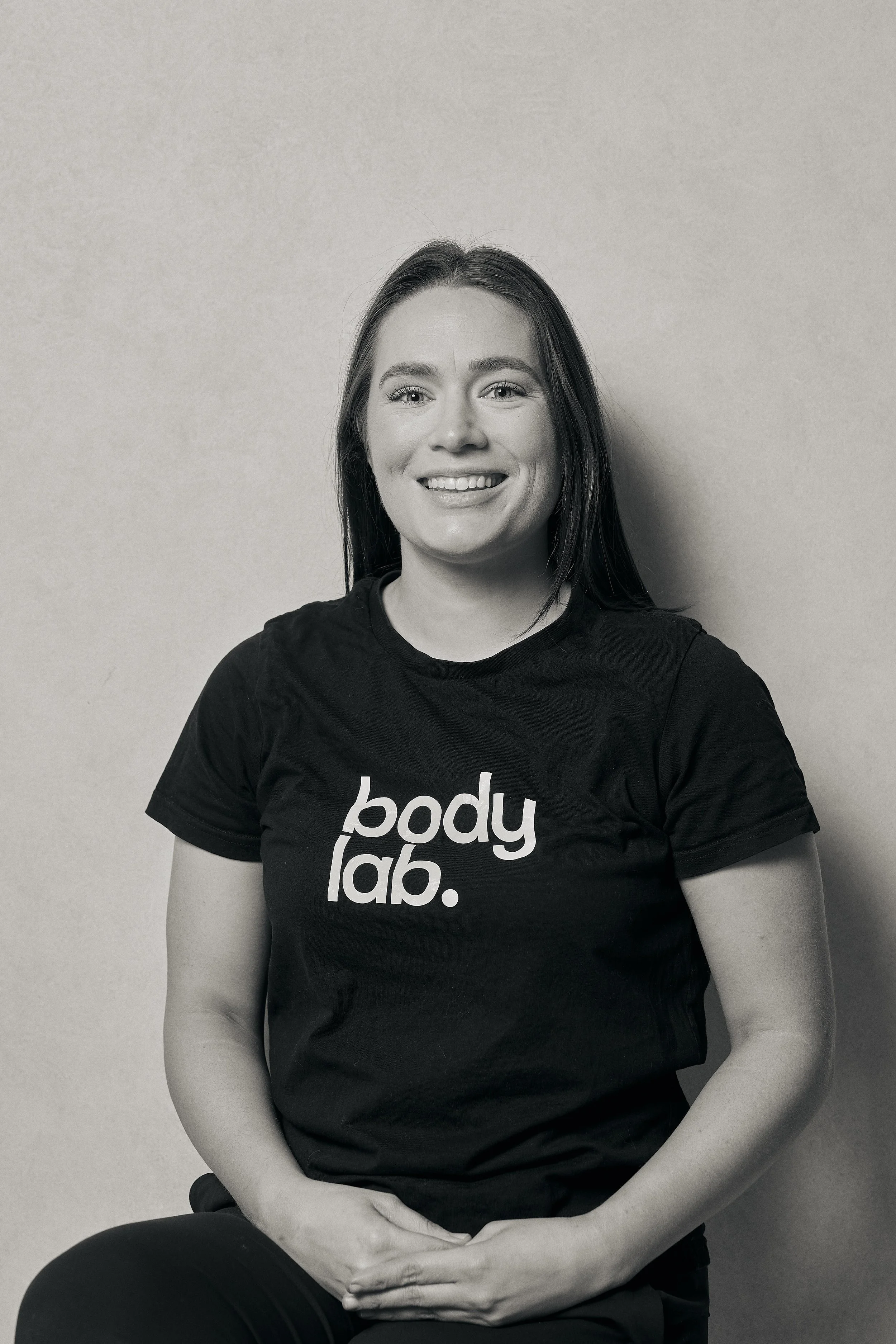 A smiling woman with long dark hair wearing a black T-shirt that says 'body lab.' poses against a plain light-colored wall.