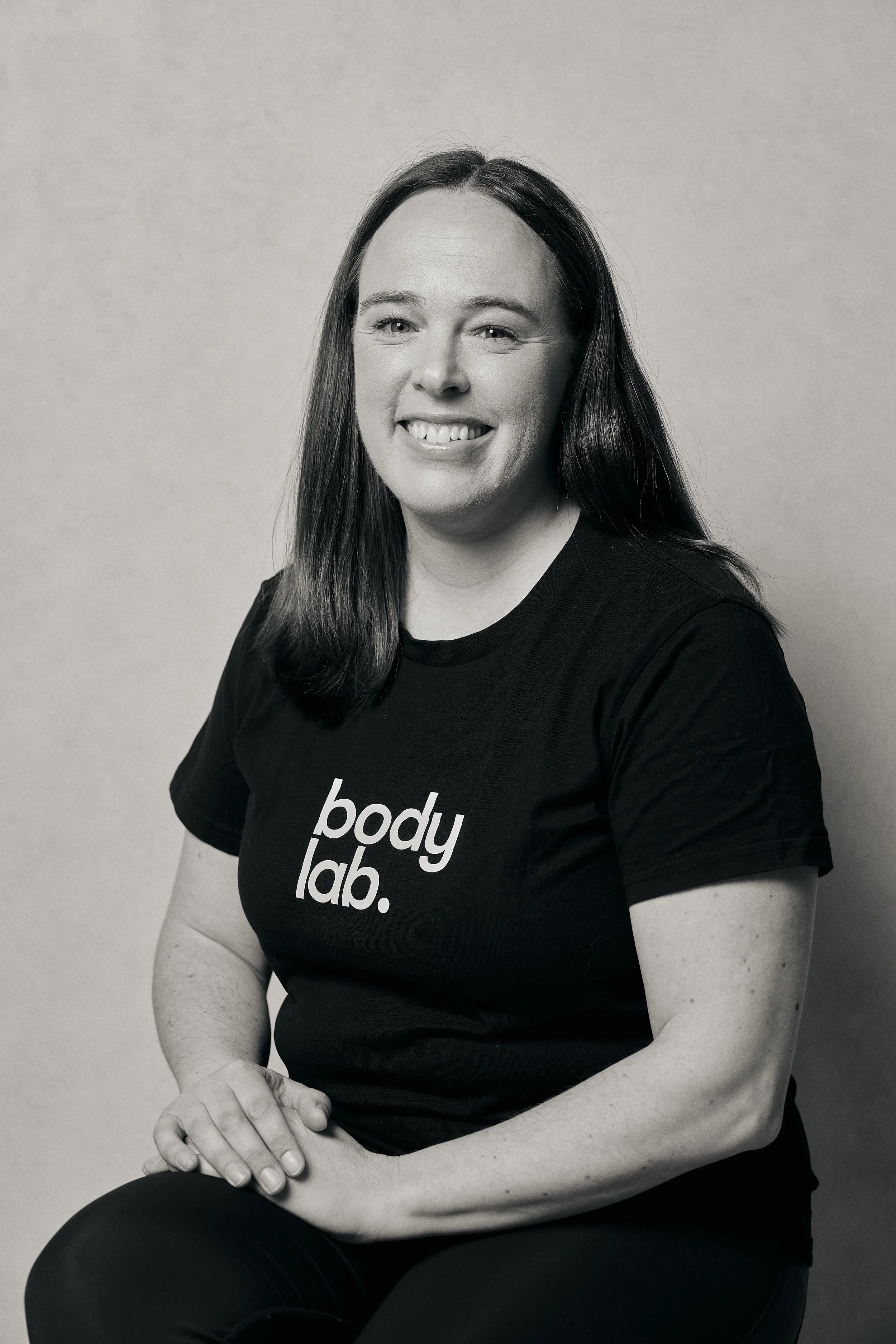 Black and white portrait of a smiling woman with long dark hair, wearing a black t-shirt with 'body lab.' printed on it, sitting with arms crossed against a plain background.