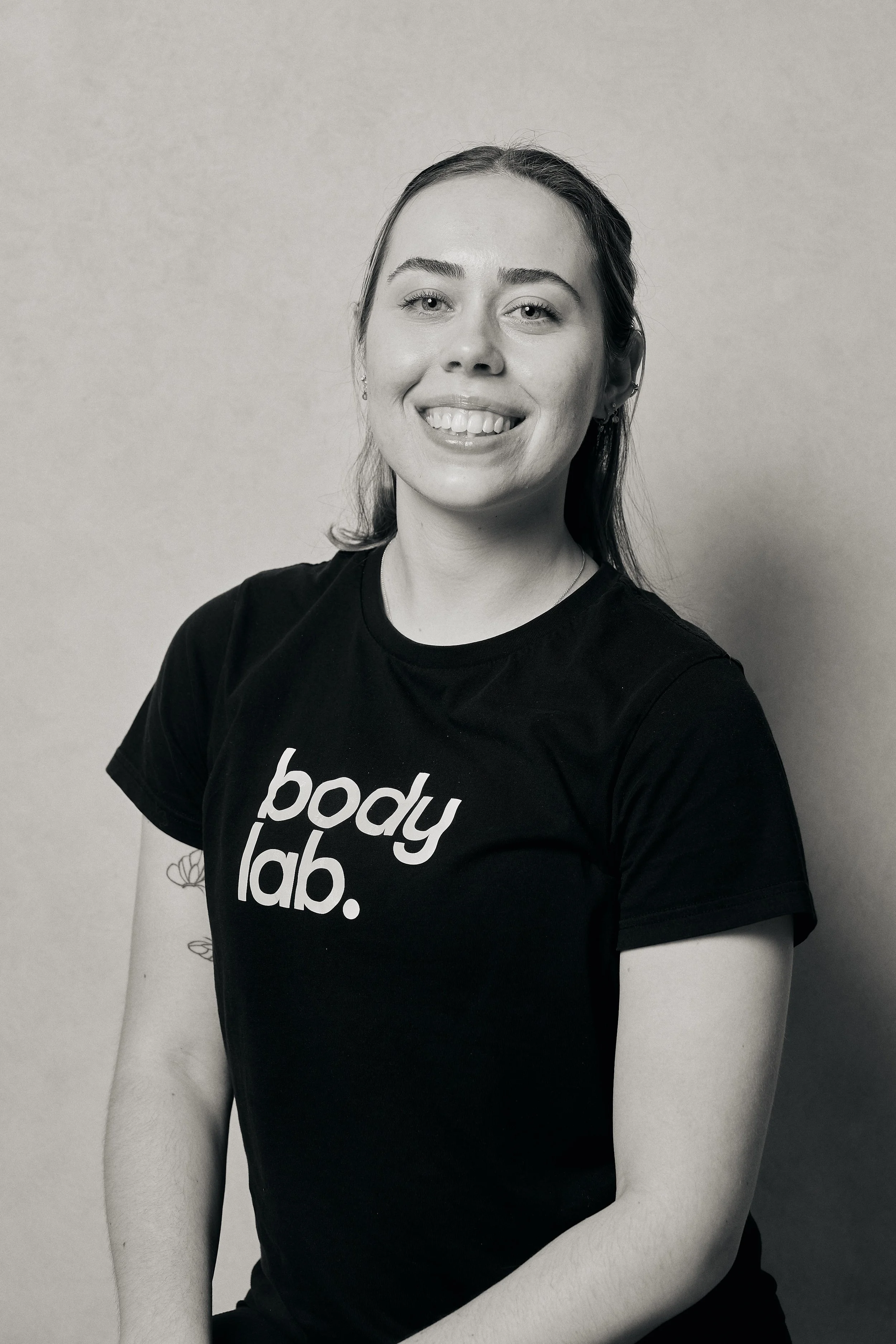 A woman smiling wearing a black t-shirt with 'body lab.' written on it, standing in front of a plain wall.
