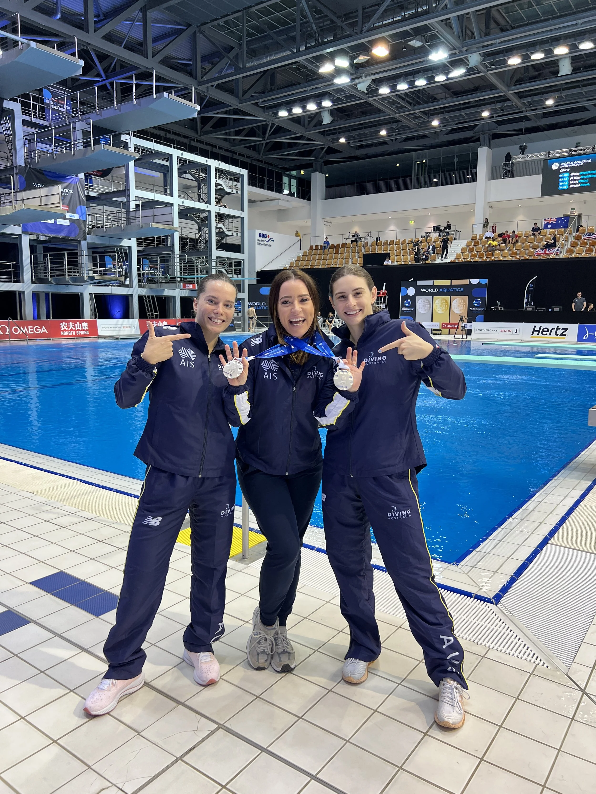 Three women in navy tracksuits standing on poolside tiles, smiling and pointing to medals they are holding. The setting is an indoor swimming pool with spectators and scoreboard in the background.