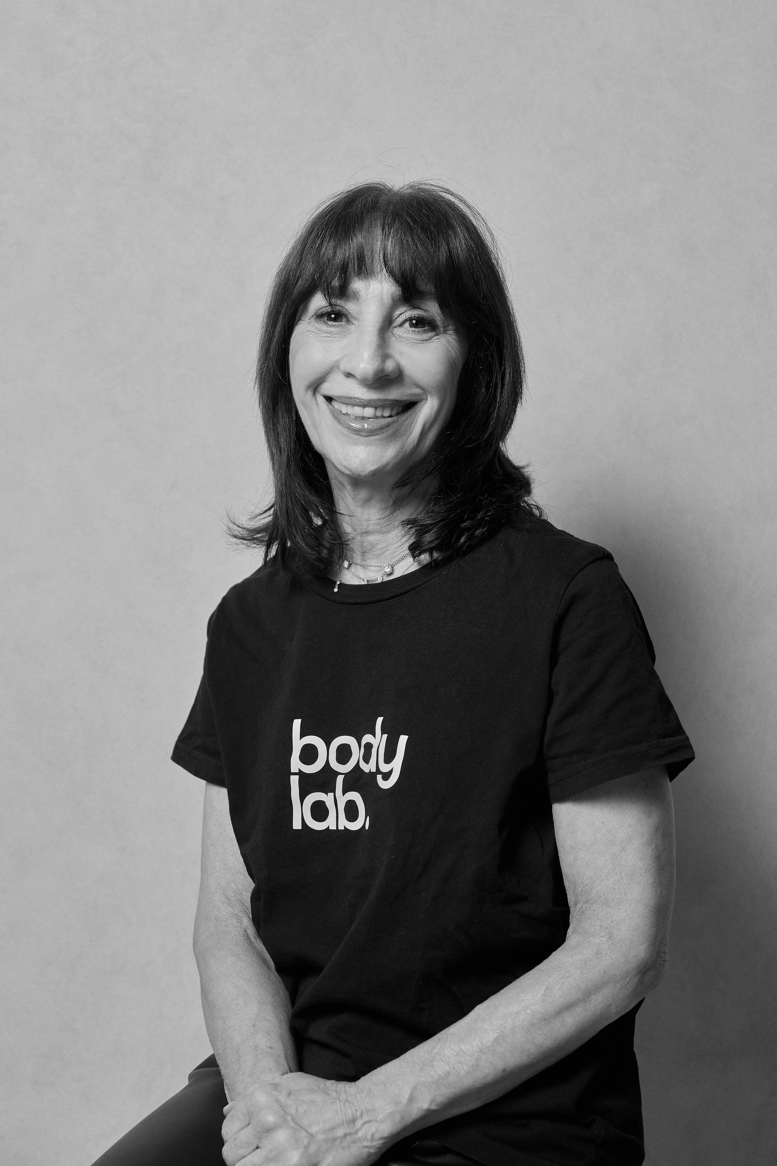 A smiling woman with shoulder-length dark hair wearing a black t-shirt with 'body lab.' printed on it, sitting against a plain background.