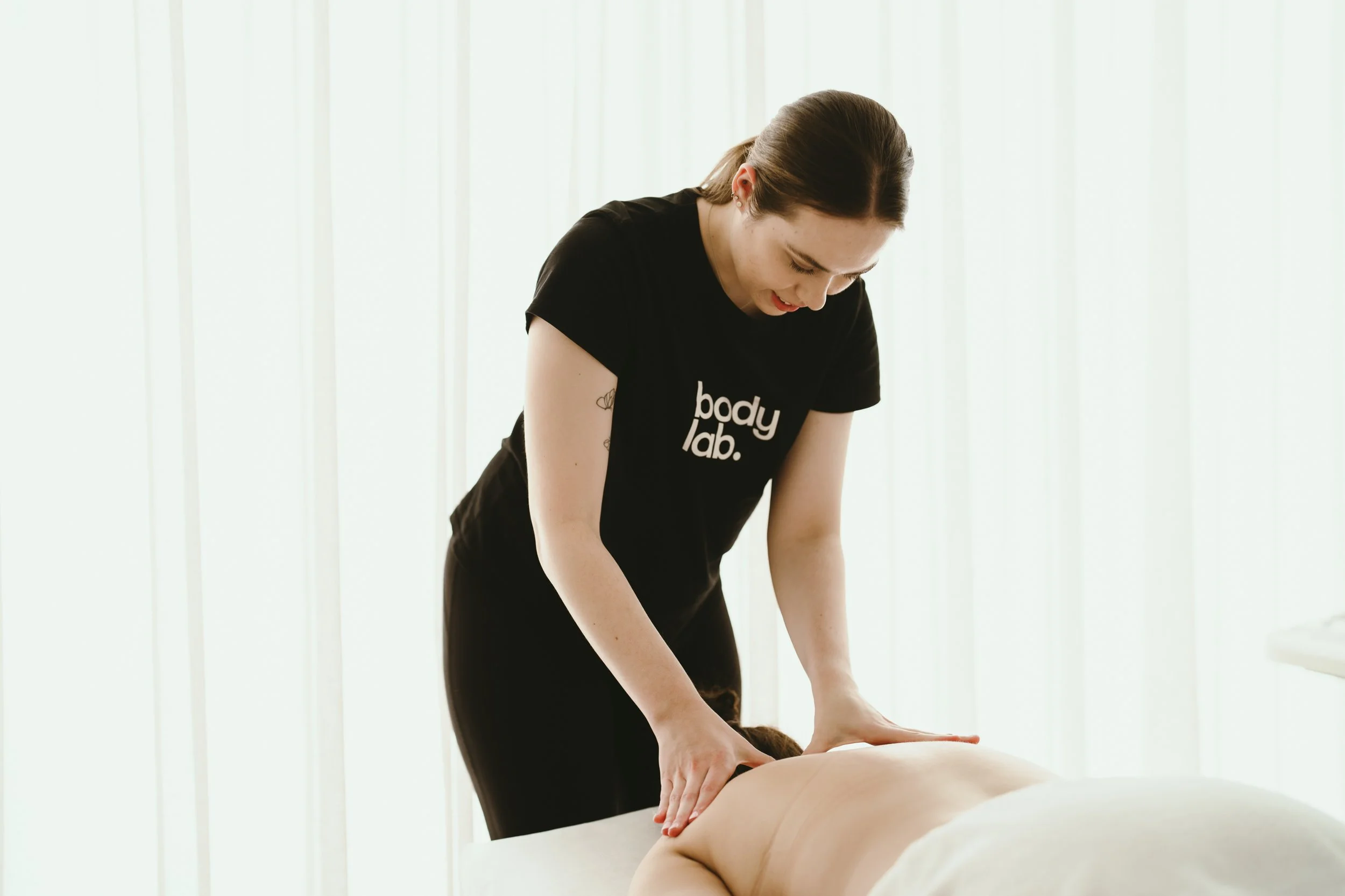 A woman giving a massage to a person lying face down on a massage table in a bright, softly lit room.