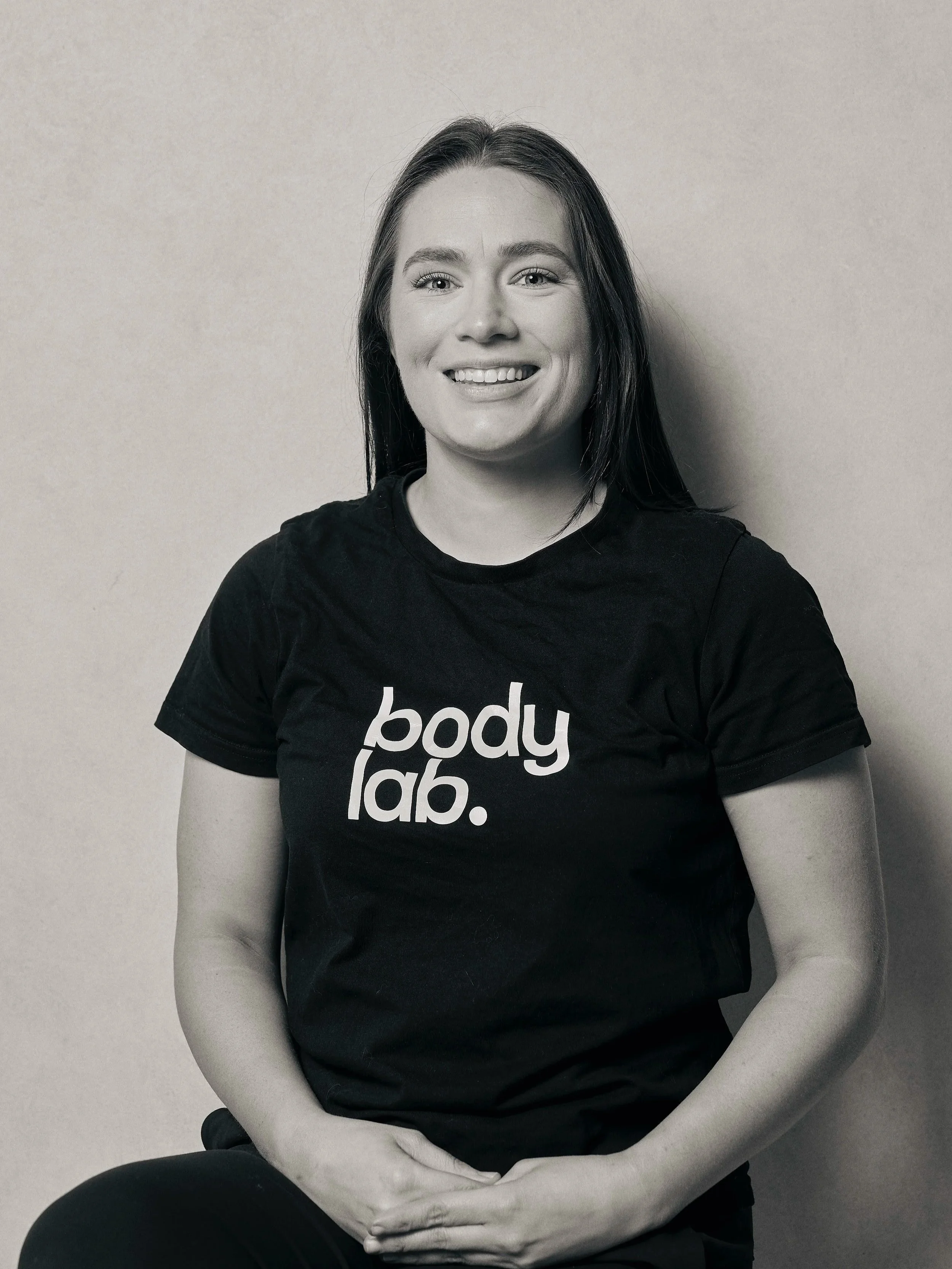 Black and white photo of a woman with long dark hair smiling, wearing a black T-shirt with "body lab." written on it, sitting against a plain wall.