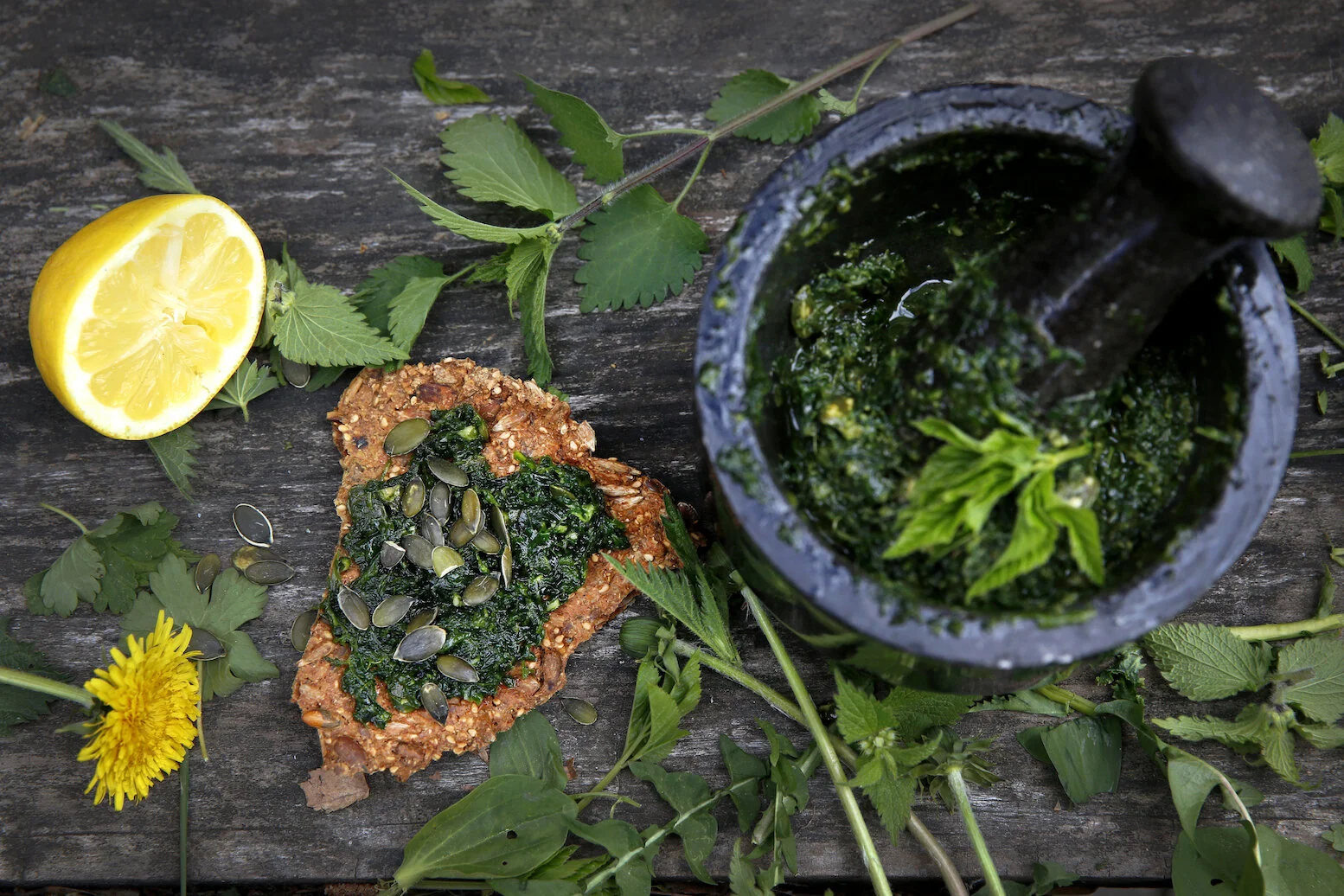 Morter med grønn ugresspest og knekkebrød med pesto og gresskarkjerner ved siden av. Ugress og en skvist sitron ligger rundt.